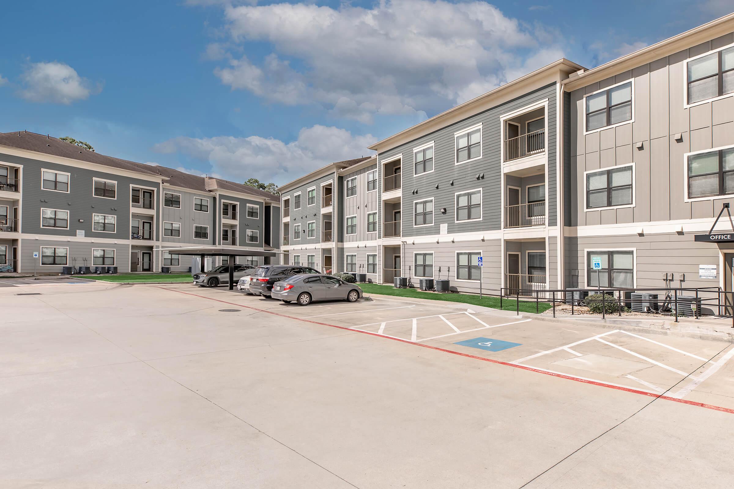 A modern apartment complex with multiple buildings. The foreground features a parking area with several parked cars and a clear sky above. The architecture includes balconies and large windows, surrounded by green grass and landscaping.