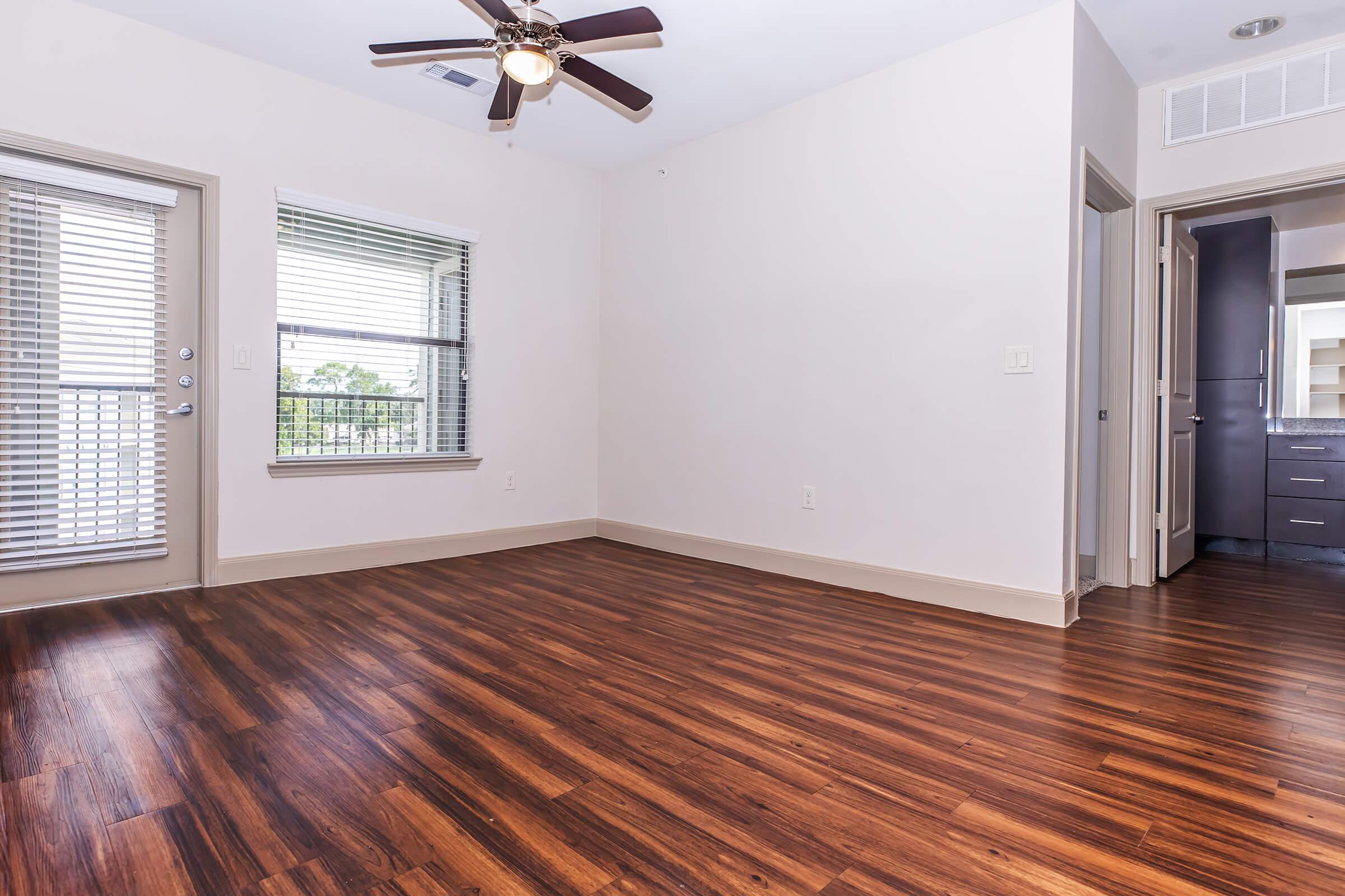 Interior of a modern living room showcasing hardwood flooring, a ceiling fan, and large windows with blinds. An open doorway leads to a bathroom, while another door provides access to a small balcony. The walls are painted a light color, creating a bright and airy atmosphere.