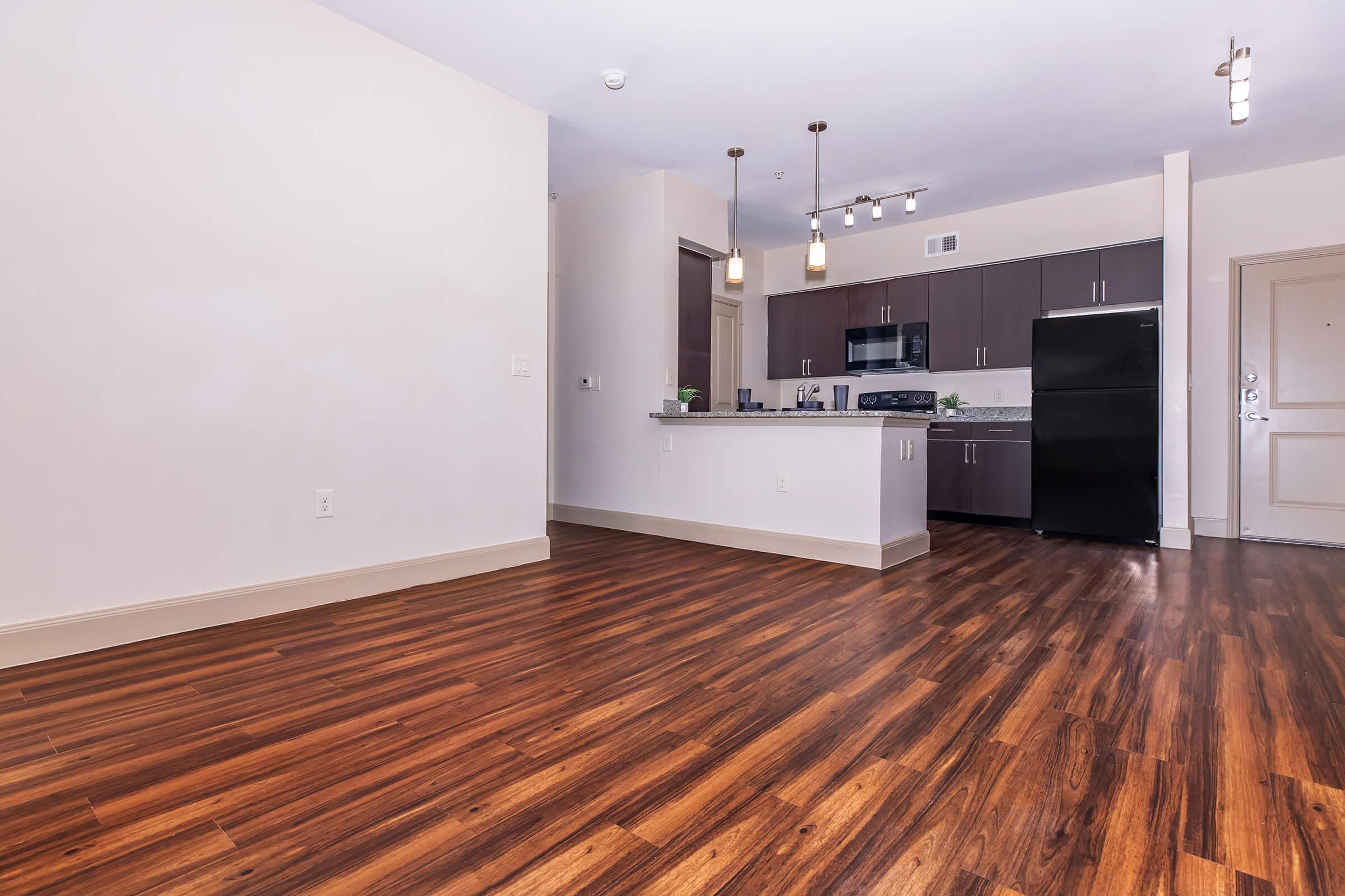 A spacious, modern apartment interior featuring an open layout with wooden flooring. The kitchen area includes dark cabinetry, stainless steel appliances, and a countertop. Natural light fills the room from a nearby window, creating a welcoming atmosphere. The entryway is visible, showcasing the contemporary design.