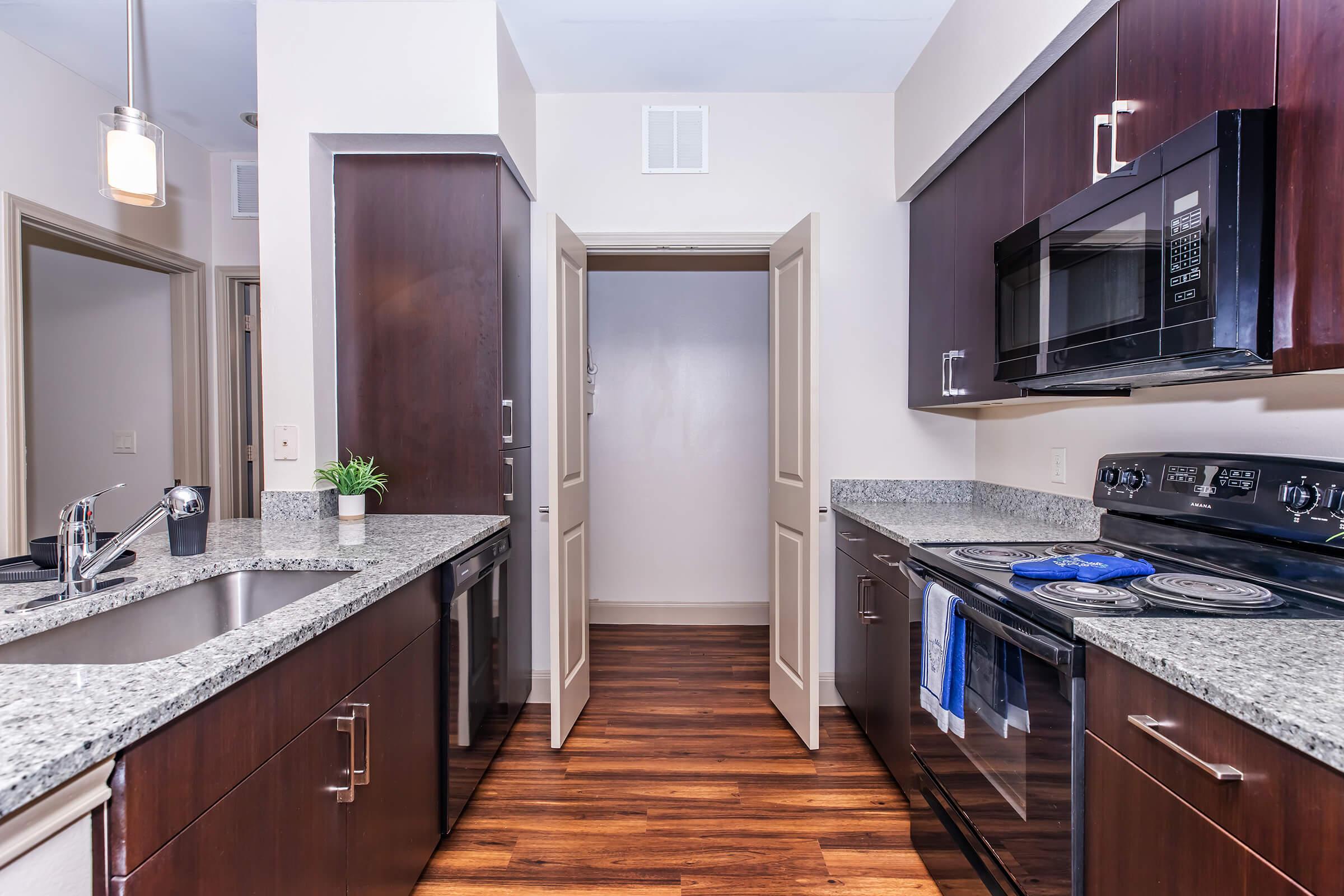 Modern kitchen featuring dark wooden cabinets and granite countertops. Includes stainless steel appliances like a microwave and oven, a double sink, and a small plant for decoration. The flooring is wooden, and there are open doors that lead to additional space. A clean and inviting atmosphere is present.