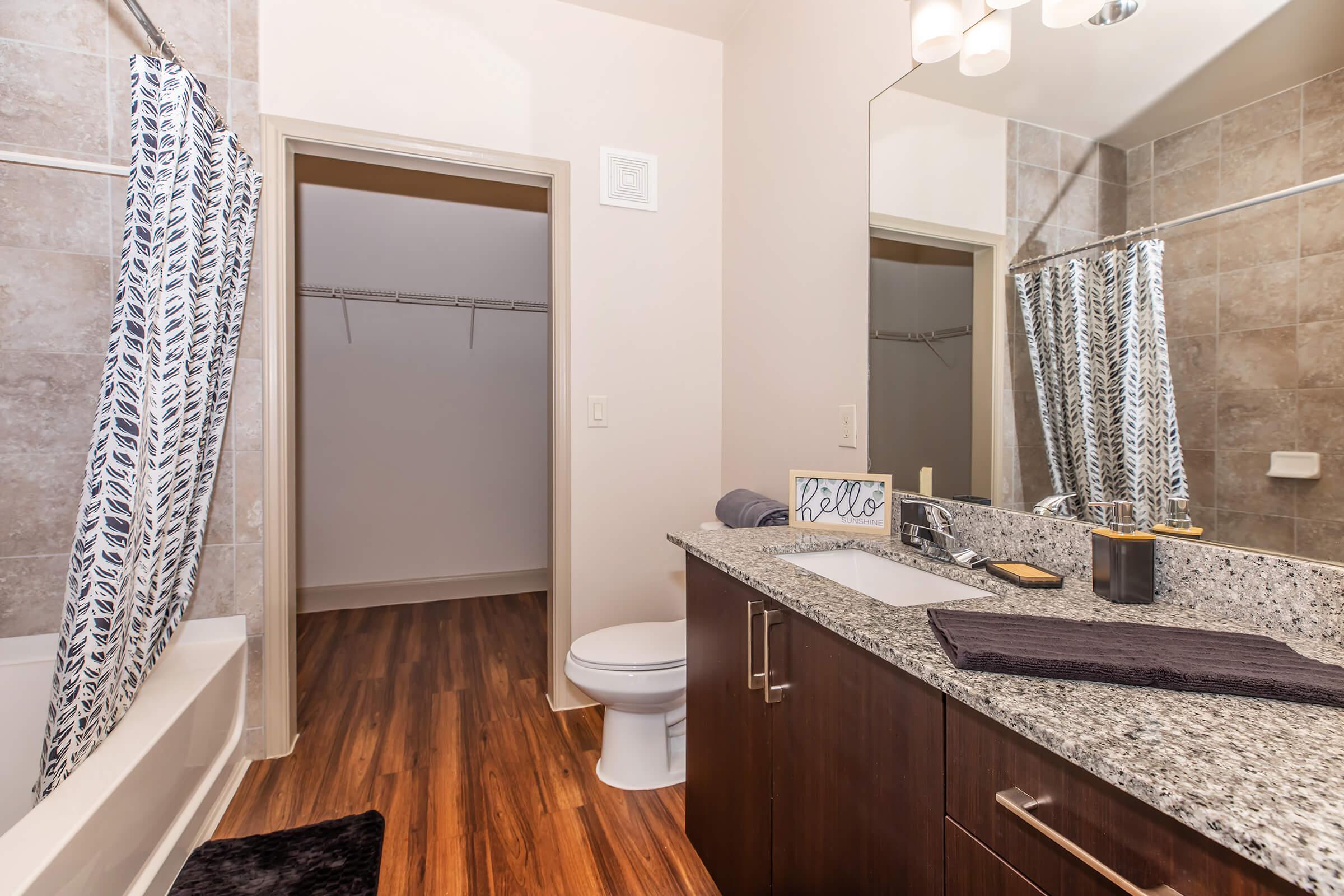 A modern bathroom featuring a vanity with a sink, dark wood cabinets, granite countertops, and a large mirror. There’s a bathtub to the left and a closet with shelves visible through an open door. The decor includes a decorative sign and a towel, with neutral-colored tiles and wooden flooring.