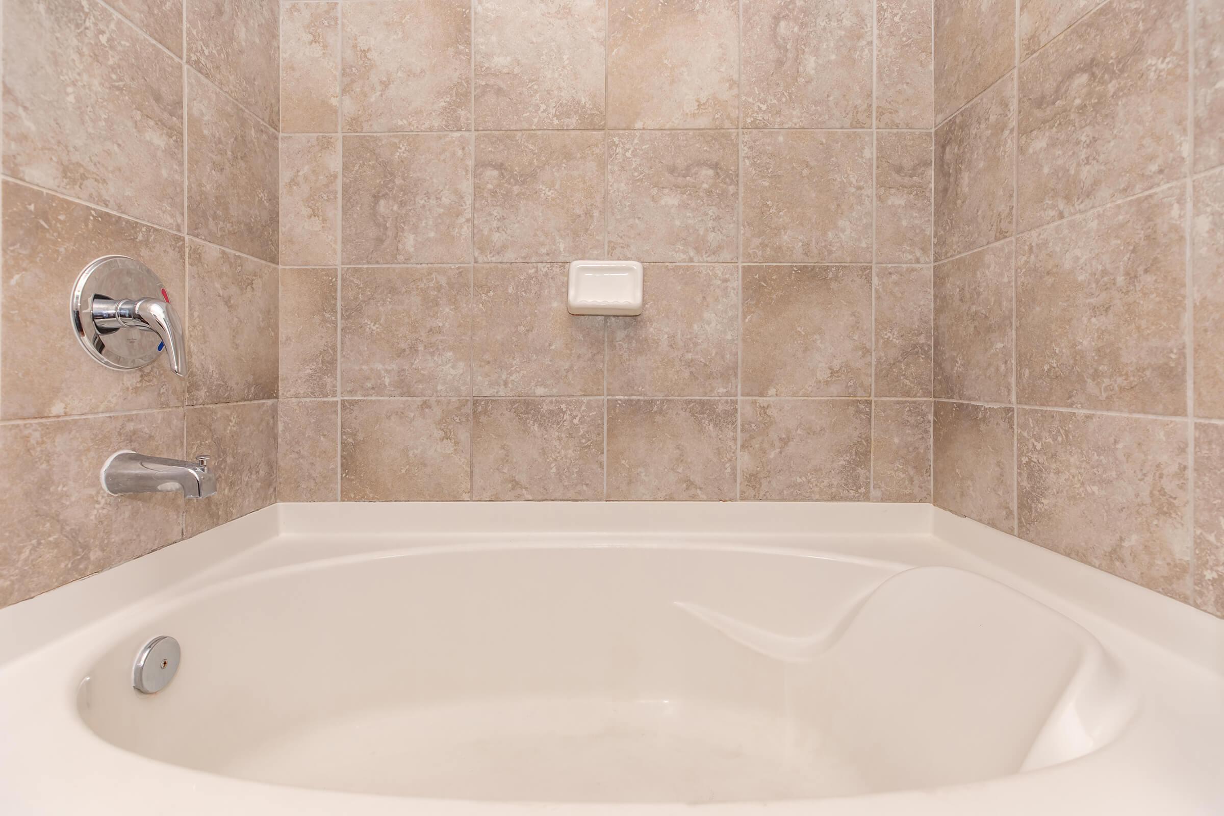 A clean, empty bathtub with a smooth white surface, set against a tiled wall featuring beige and light brown squares. The bathtub has a faucet on one side and a soap holder on the wall. The overall atmosphere is calm and minimalist, suitable for relaxation.