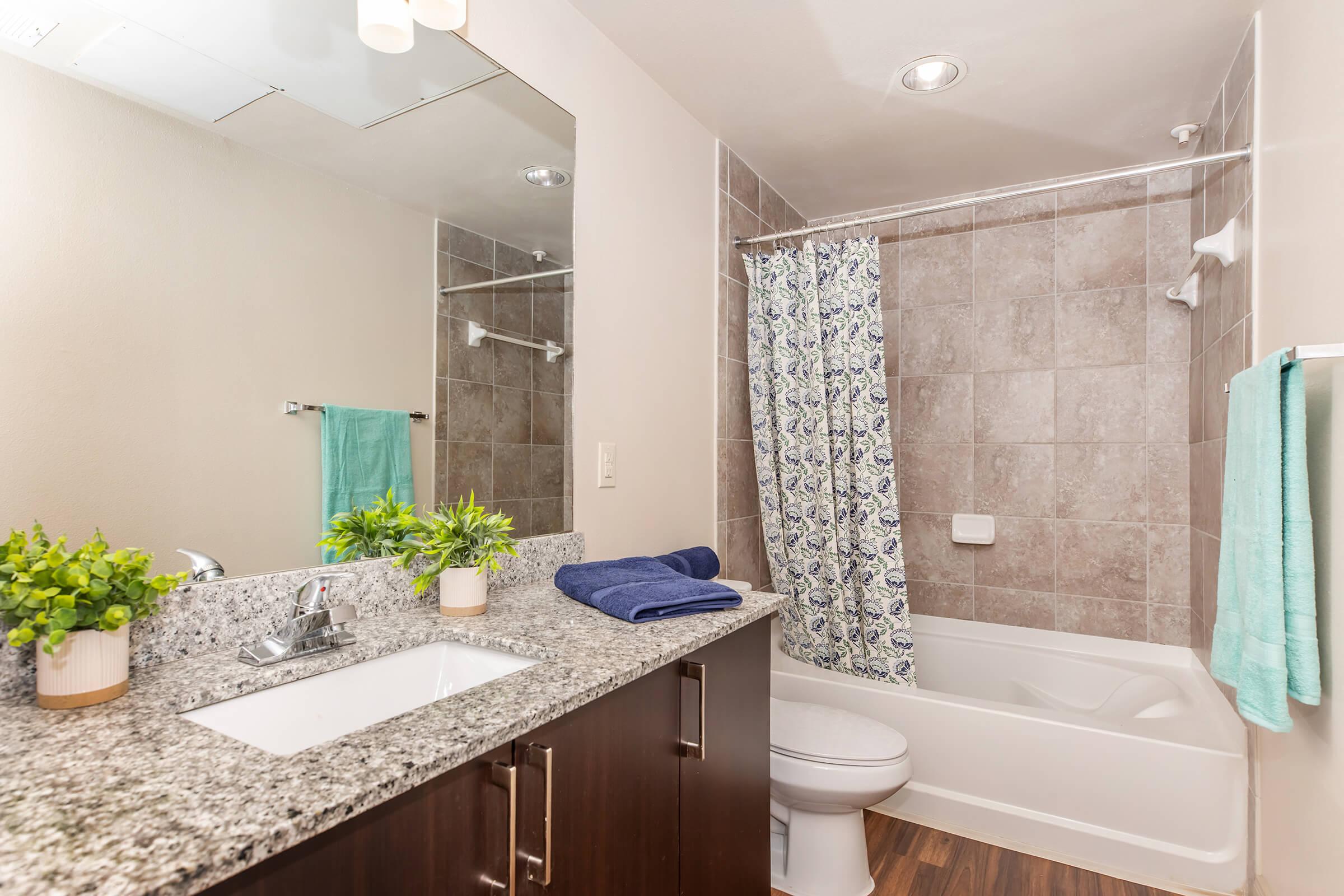 A modern bathroom featuring a granite countertop with a sink, a large mirror, and decorative plants. There is a bathtub with a shower curtain in a floral pattern, a toilet, and a towel rack with a turquoise towel. The walls are light-colored, and the floor is made of wood-like tiles.