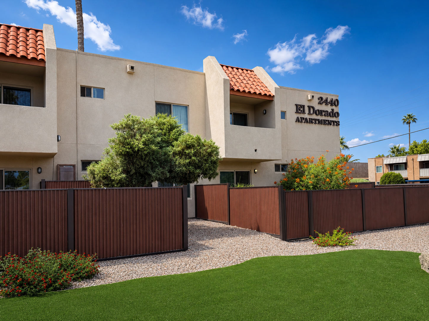 A view of the El Dorado Apartments building with a light beige exterior and a red-tiled roof. The entrance is bordered by shrubs and a wooden fence, and there is manicured green grass in the foreground. The sky is clear with a few clouds.