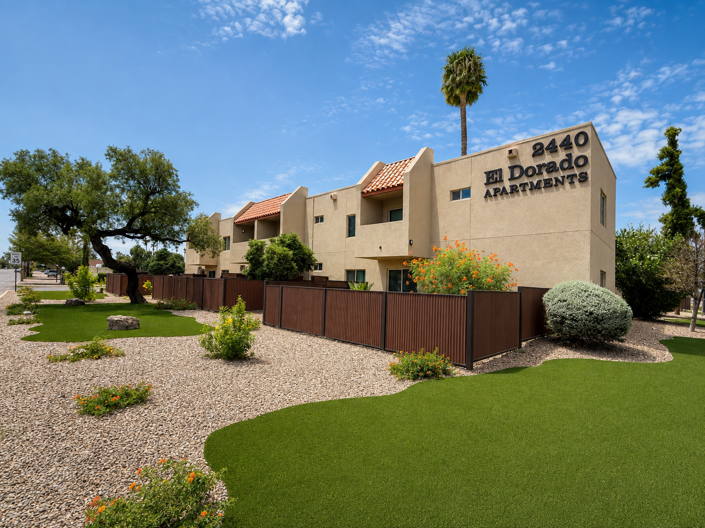 A sunny view of the El Dorado Apartments, featuring a modern, two-story building with a terracotta roof. The landscape includes green grass, desert plants, and decorative stone pathways. A palm tree is visible, and there's a clear blue sky overhead, creating a welcoming atmosphere.