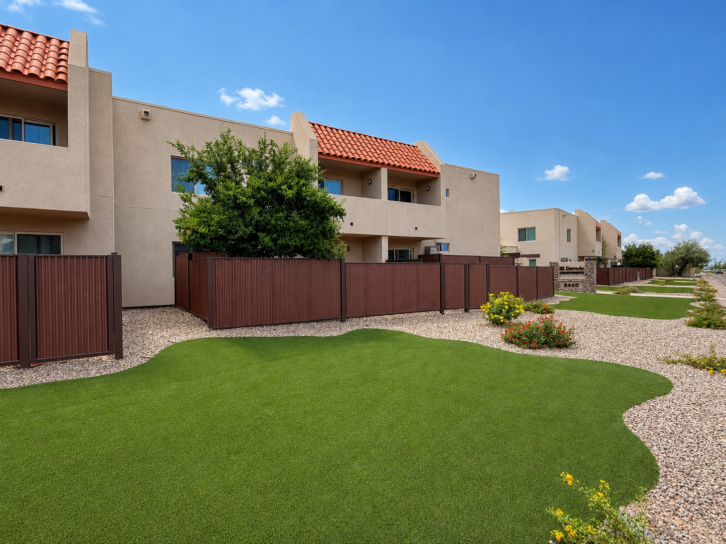 A view of a well-maintained apartment complex featuring multiple buildings with tan stucco exteriors and red tile roofs. Neatly trimmed green lawns are bordered by gravel paths and wooden fences, with a few flowering bushes visible in the landscape under a sunny blue sky and scattered clouds.