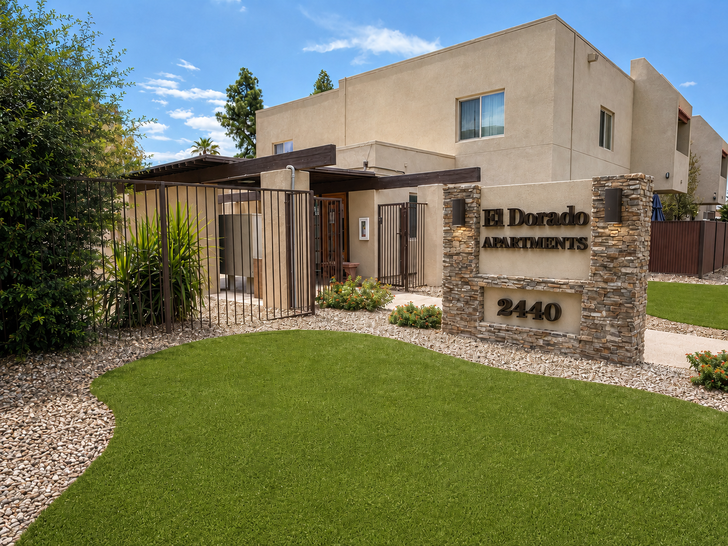 A modern apartment building entrance featuring a sign that reads "El Dorado Apartments." The exterior includes landscaped greenery, decorative stonework, and well-maintained paths, with a clear blue sky in the background, creating a welcoming atmosphere.