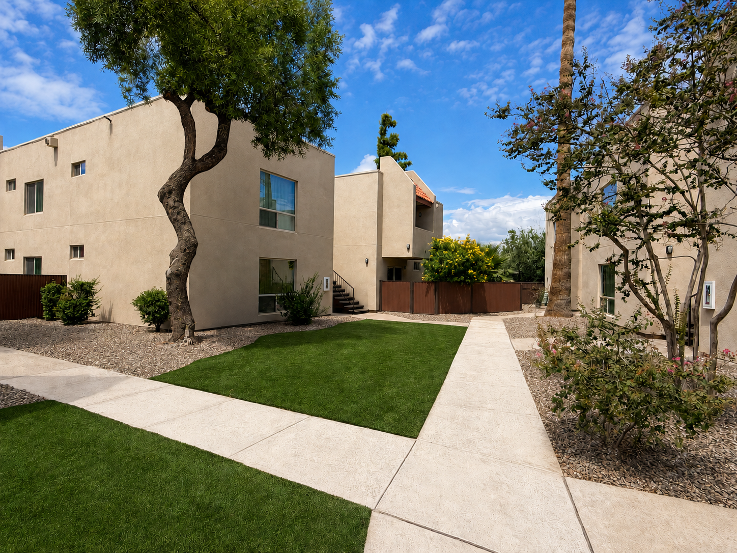 A landscaped area featuring two beige apartment buildings surrounded by green lawns. There are concrete walkways leading between the buildings, with shrubs and trees adding to the greenery. The sky is partly cloudy, creating a bright and inviting atmosphere.