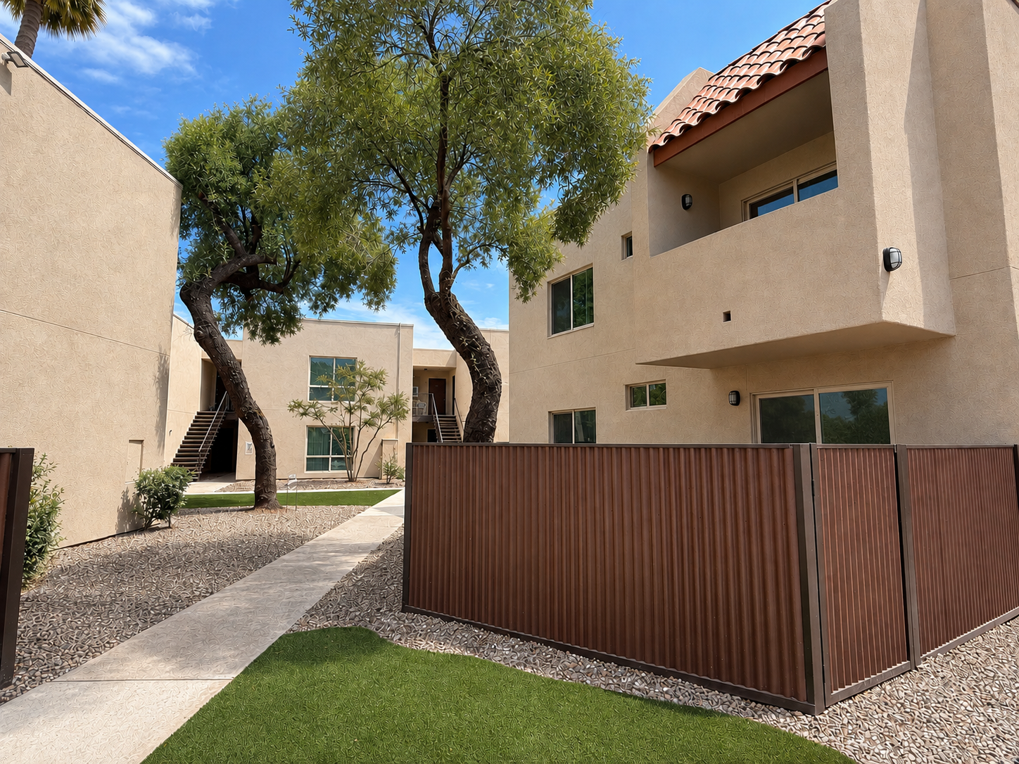 View of a residential area featuring beige buildings with tiled rooftops, surrounded by greenery. There are two trees, a pathway, and a brown fence separating the grassy area from the buildings. The sky is clear with a few clouds, creating a bright and inviting atmosphere.