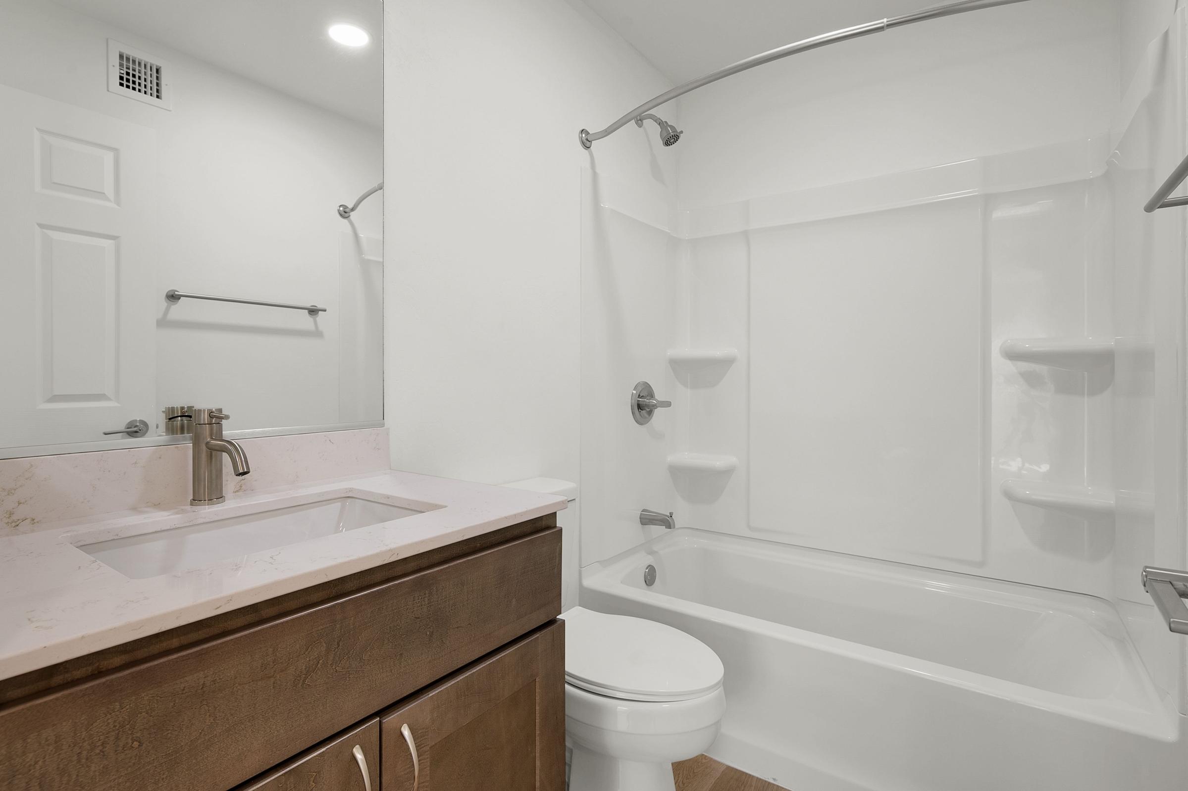 A modern bathroom featuring a shower-tub combination with a white wall and gray tile flooring. There is a wooden vanity with a sink, a mirror above it, and a towel bar on the wall. The lighting is bright, and the space appears clean and well-maintained.