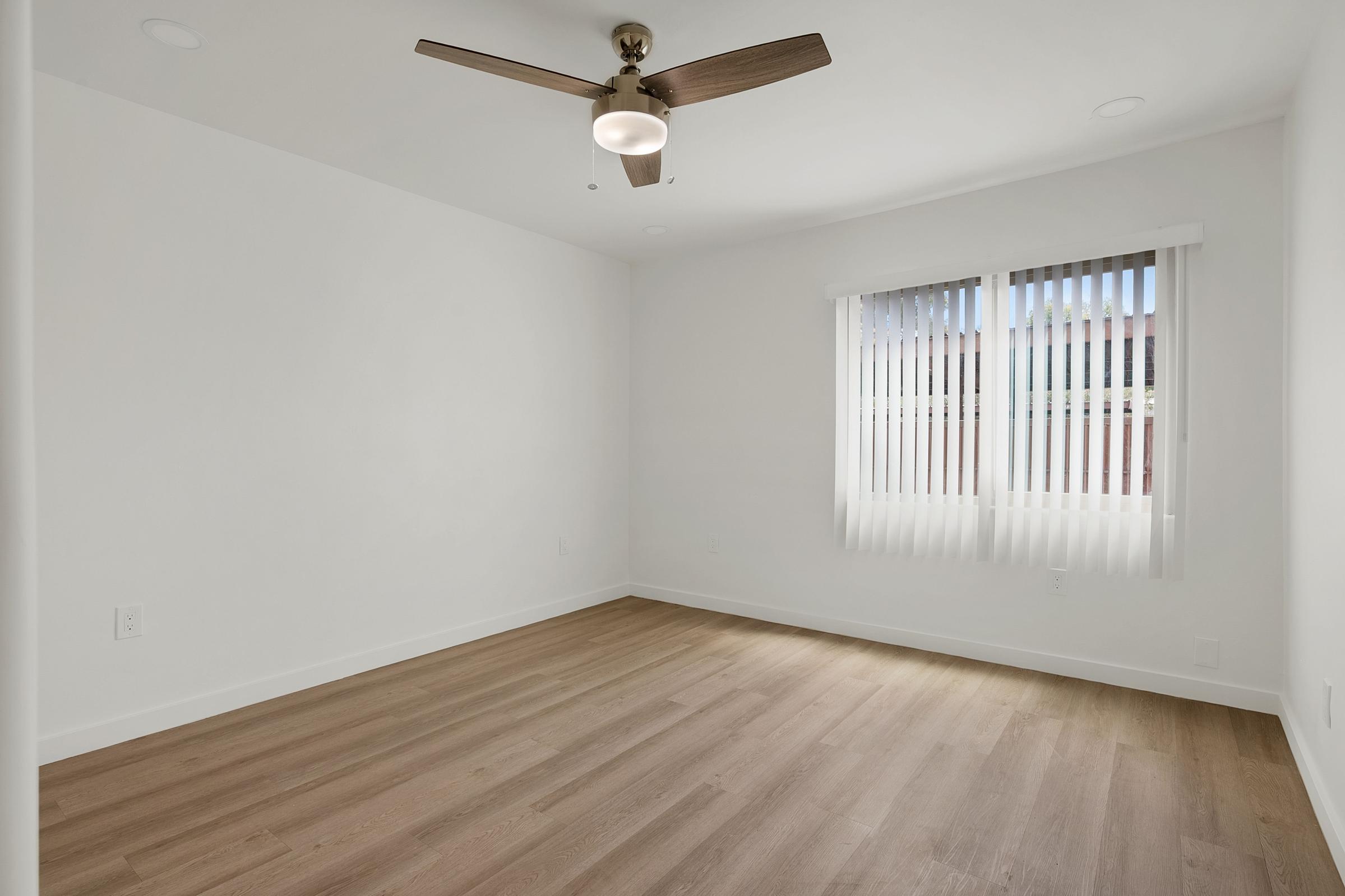 Empty room with light-colored walls and wooden flooring. A ceiling fan is centered above, and there are two windows with vertical blinds allowing natural light to enter. The room appears modern and unfurnished, creating a minimalist aesthetic.