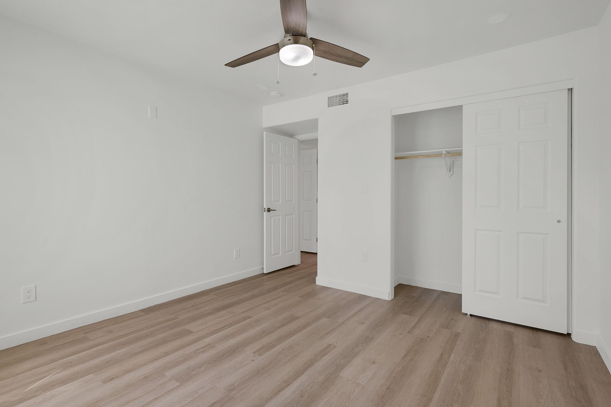 A clean, empty room featuring light-colored walls and a wood-style floor. There is a ceiling fan with three blades, and a closet with sliding doors is visible on the right. The room has a doorway leading to another area, enhancing the sense of spaciousness. Overall, the space feels bright and airy.