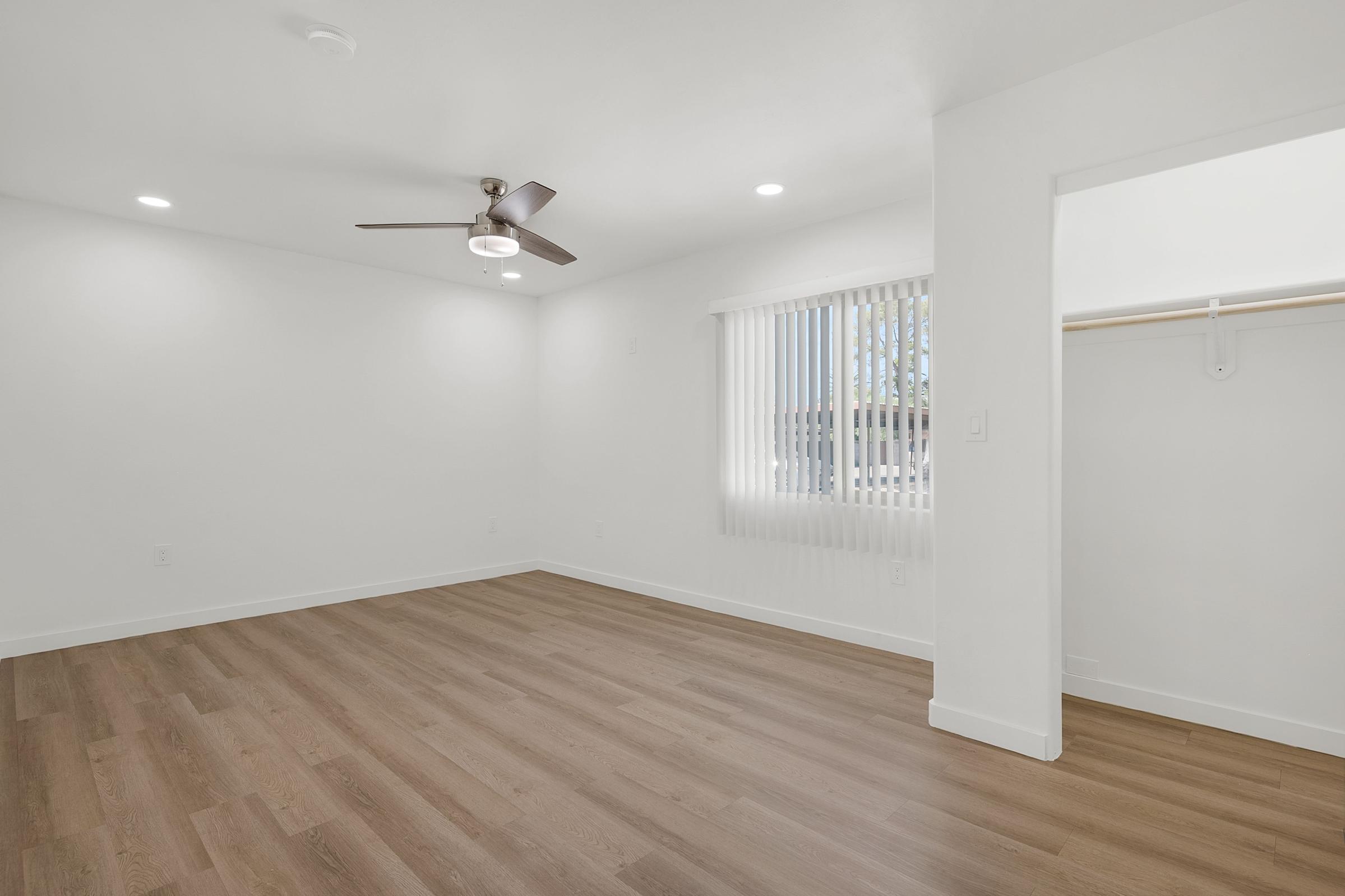 A bright, unfurnished room with white walls and hardwood-style flooring. It features a ceiling fan, recessed lighting, and a large window with vertical blinds. A closet is visible on the right side of the image, contributing to the room's spacious feel.