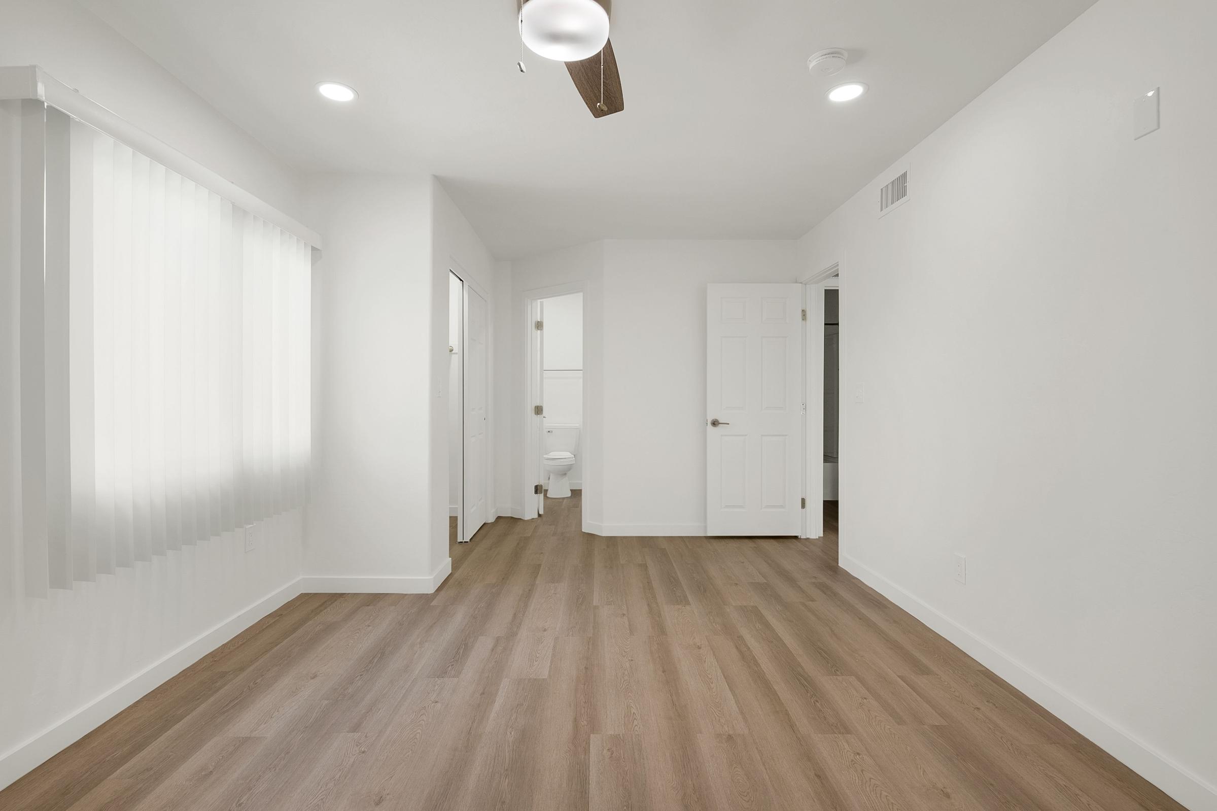 A bright, empty room with light-colored walls and wooden flooring. There is a ceiling fan and natural light coming in from a window with blinds. A doorway leads to a bathroom in the background, visible through an open door. The space is clean and modern, creating a minimalistic feel.