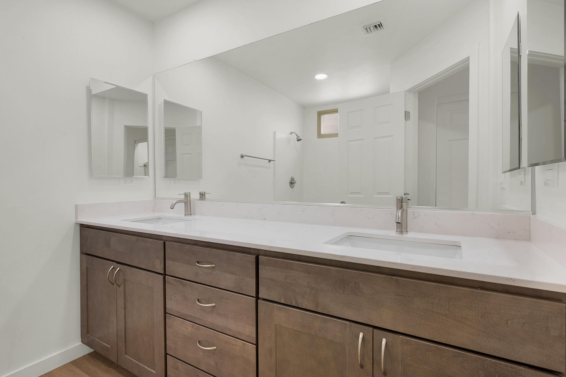 A modern bathroom featuring a double-sink vanity with a light-colored countertop, two mirrors above the sinks, and wooden cabinetry. The space is well-lit with natural and artificial lighting, and a shower area is visible in the background, along with a door leading to another room.