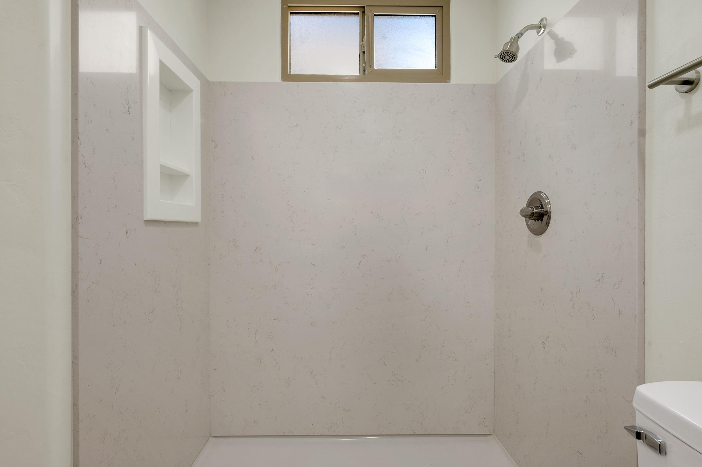 A clean, modern shower area featuring light-colored stone walls, a chrome showerhead, and a built-in niche for toiletries. A small window at the top provides natural light. The space is simple and minimalist, with a white shower base and a towel rack on the side.