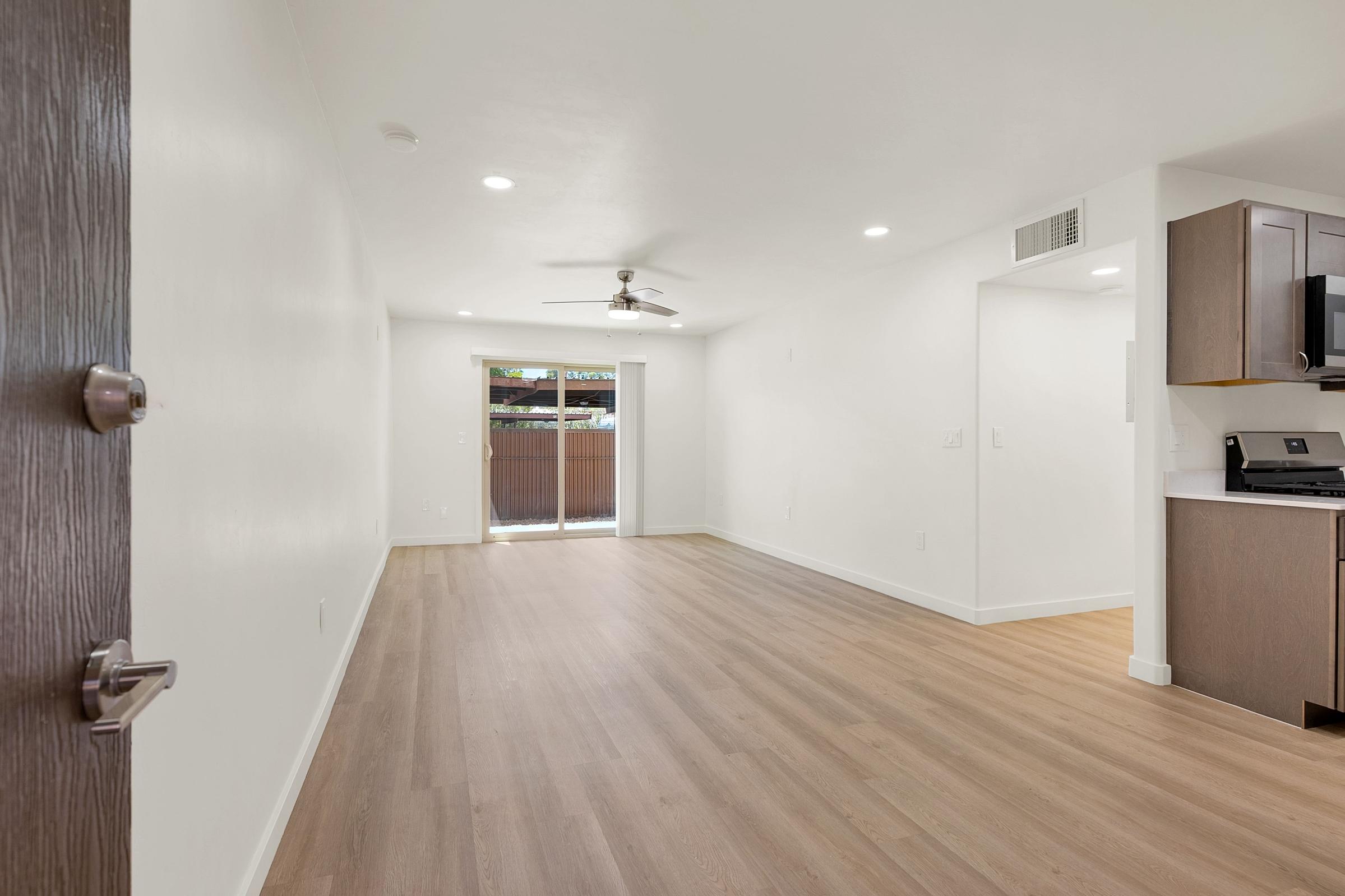 Bright and spacious living area featuring modern light hardwood flooring, fresh white walls, and a ceiling fan. There is an open layout with a view of a patio door leading to an outdoor space, and a kitchen area with contemporary cabinetry and appliances visible in the corner.