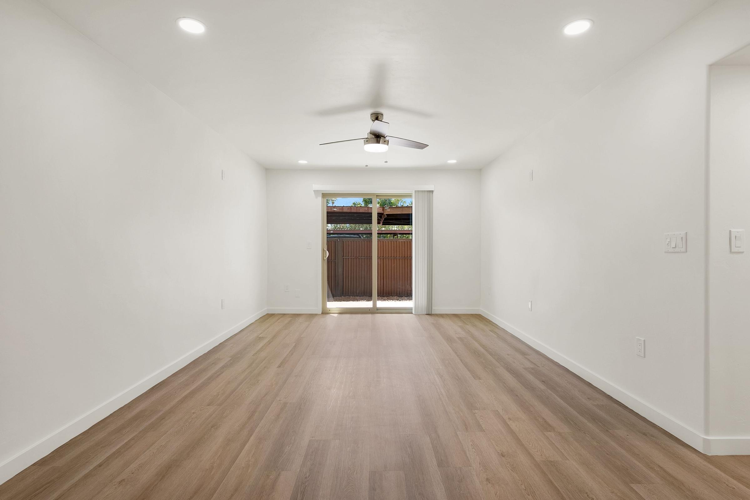A bright, empty room with light-colored walls and a ceiling fan. The space features hardwood flooring and a large sliding door leading to a fenced outdoor area, allowing natural light to fill the room. The minimalistic design creates an airy and open atmosphere.