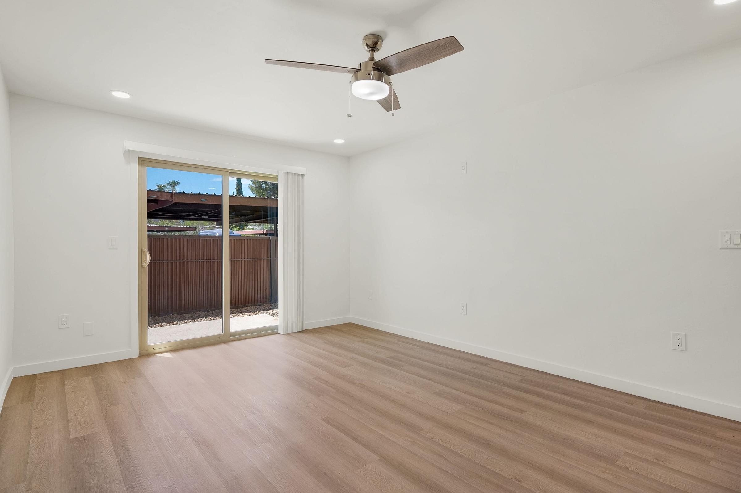 A bright, empty room with light-colored walls and a wooden floor. A ceiling fan is installed above, and there is a sliding glass door that opens to an outdoor area with a fence. Natural light streams in through the door, creating a spacious and airy atmosphere.