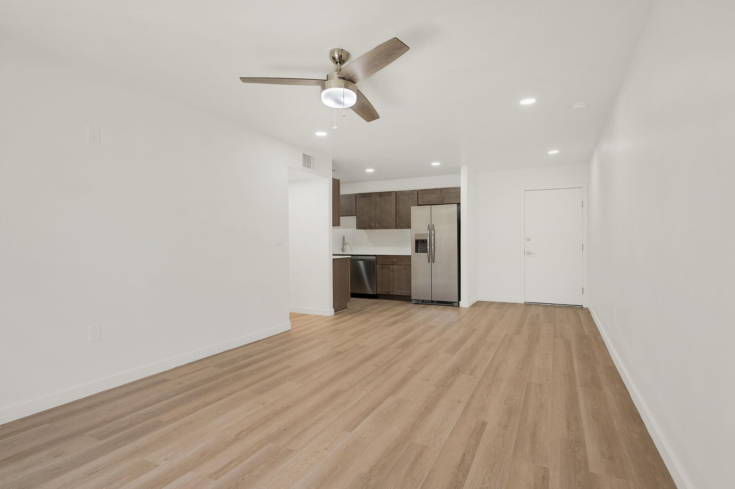 A spacious, empty living area with light wood flooring, a ceiling fan, and white walls. On one side, there is a small kitchen featuring dark wood cabinets and stainless steel appliances. Natural light floods the room, enhancing its open and airy feel. The opposite wall has a door leading outside.