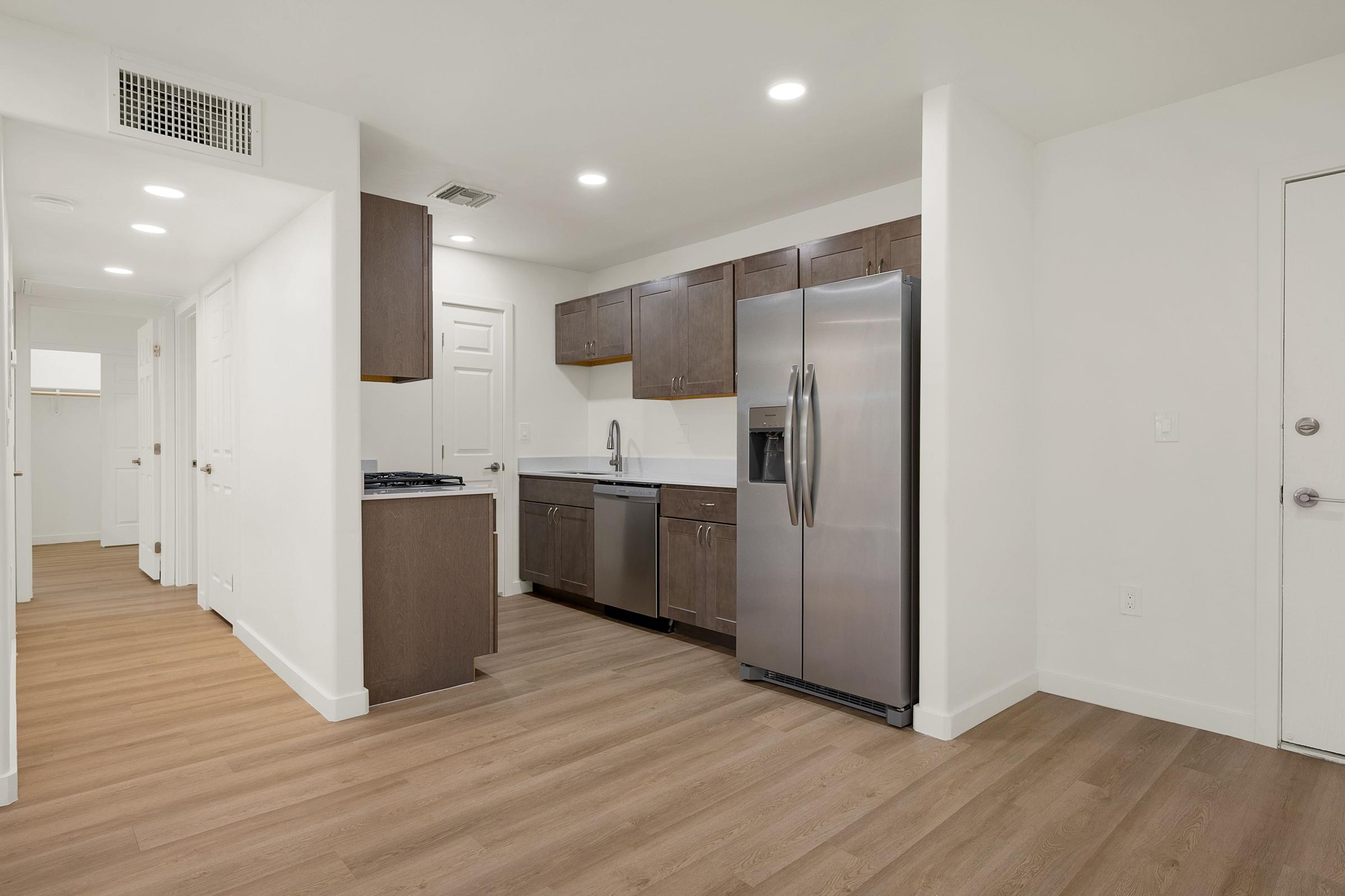 A modern kitchen featuring stainless steel appliances, including a refrigerator and dishwasher. The cabinets are dark wood, contrasting with the light walls and flooring. The space is bright, with recessed lighting and an open layout leading to a hallway.
