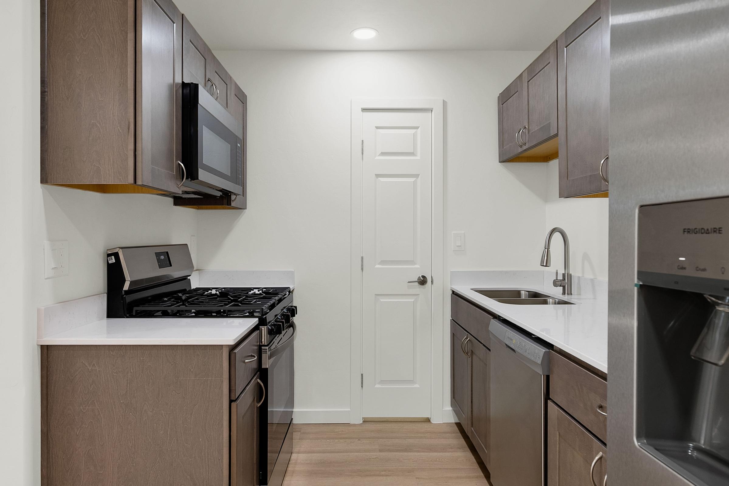 Modern kitchen featuring gray cabinets, stainless steel appliances including a gas stove, microwave, and refrigerator with ice dispenser. The countertops are light-colored, and there is a single-basin sink with a sleek faucet. A door leads to another room, and the floor is light wood. Well-lit with overhead lighting.
