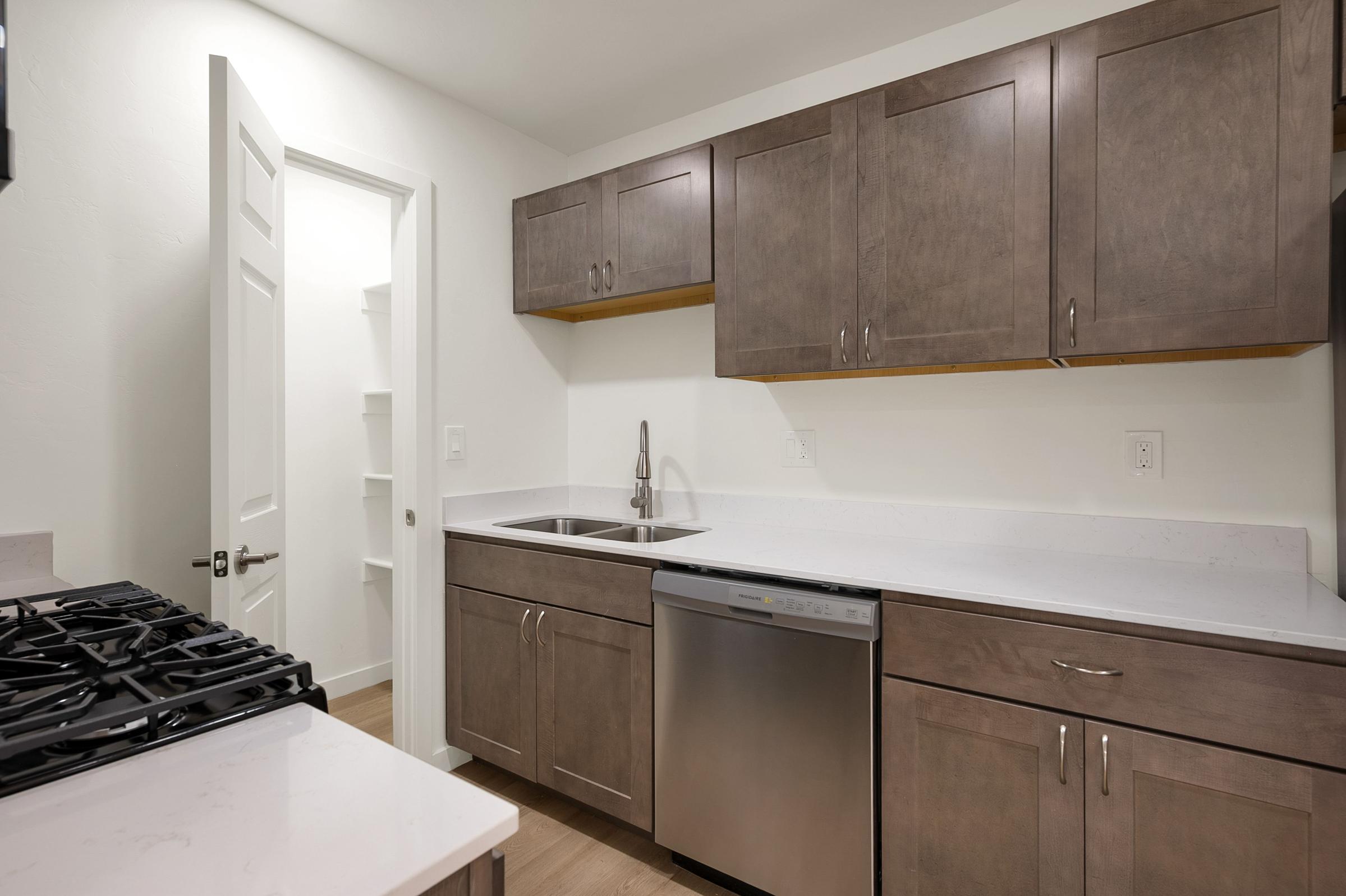 Modern kitchen featuring light-colored cabinets, a stainless steel dishwasher, under-mounted sink, and a gas stove. The countertop is white, and there's a door leading to a small pantry area. The space is well-lit and designed with a contemporary aesthetic.