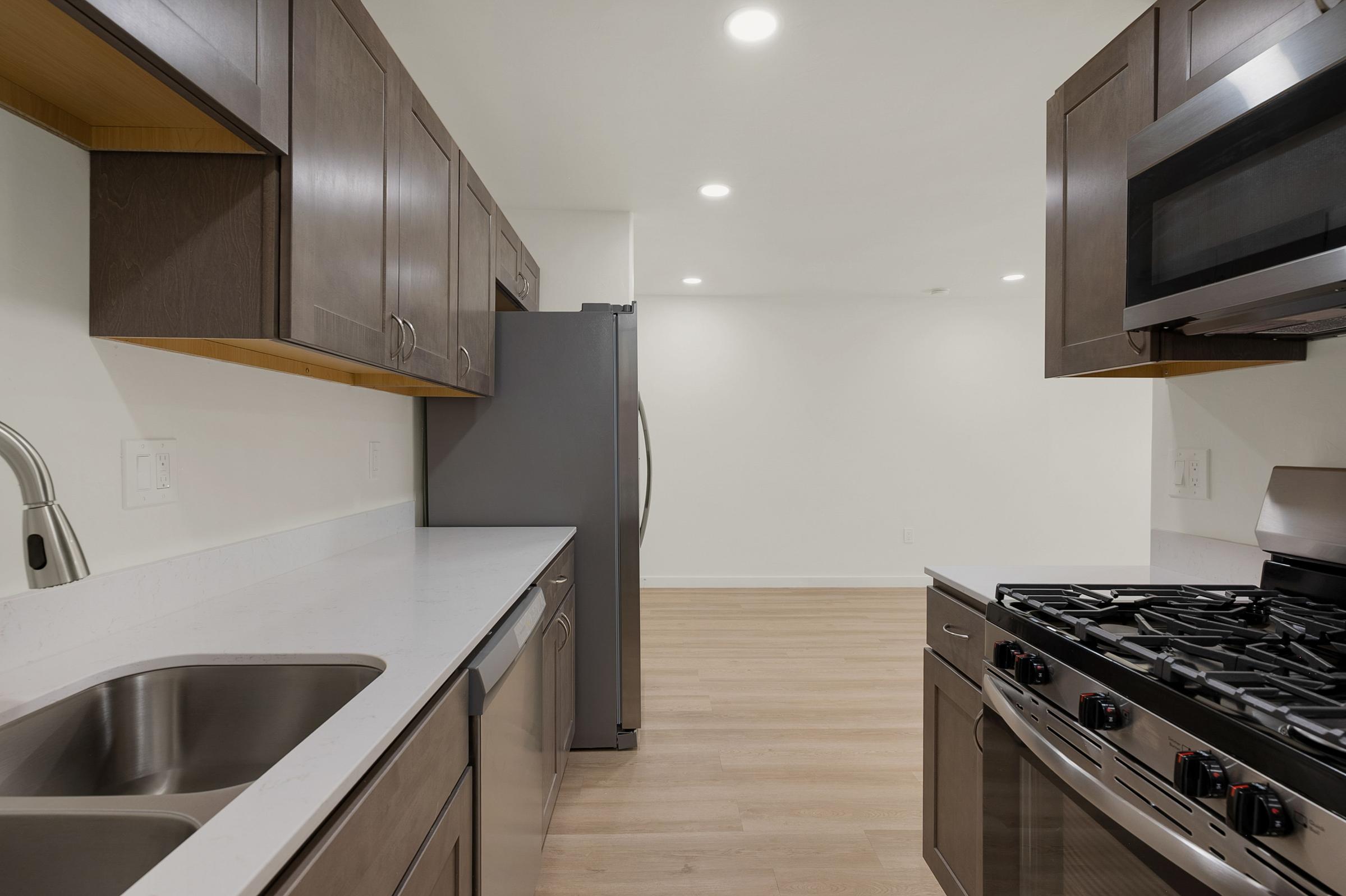A modern kitchen featuring dark wood cabinetry, stainless steel appliances, including a refrigerator and a gas stove, and a white countertop. The space has a bright, neutral-toned wall and wooden flooring, creating a clean and inviting atmosphere.