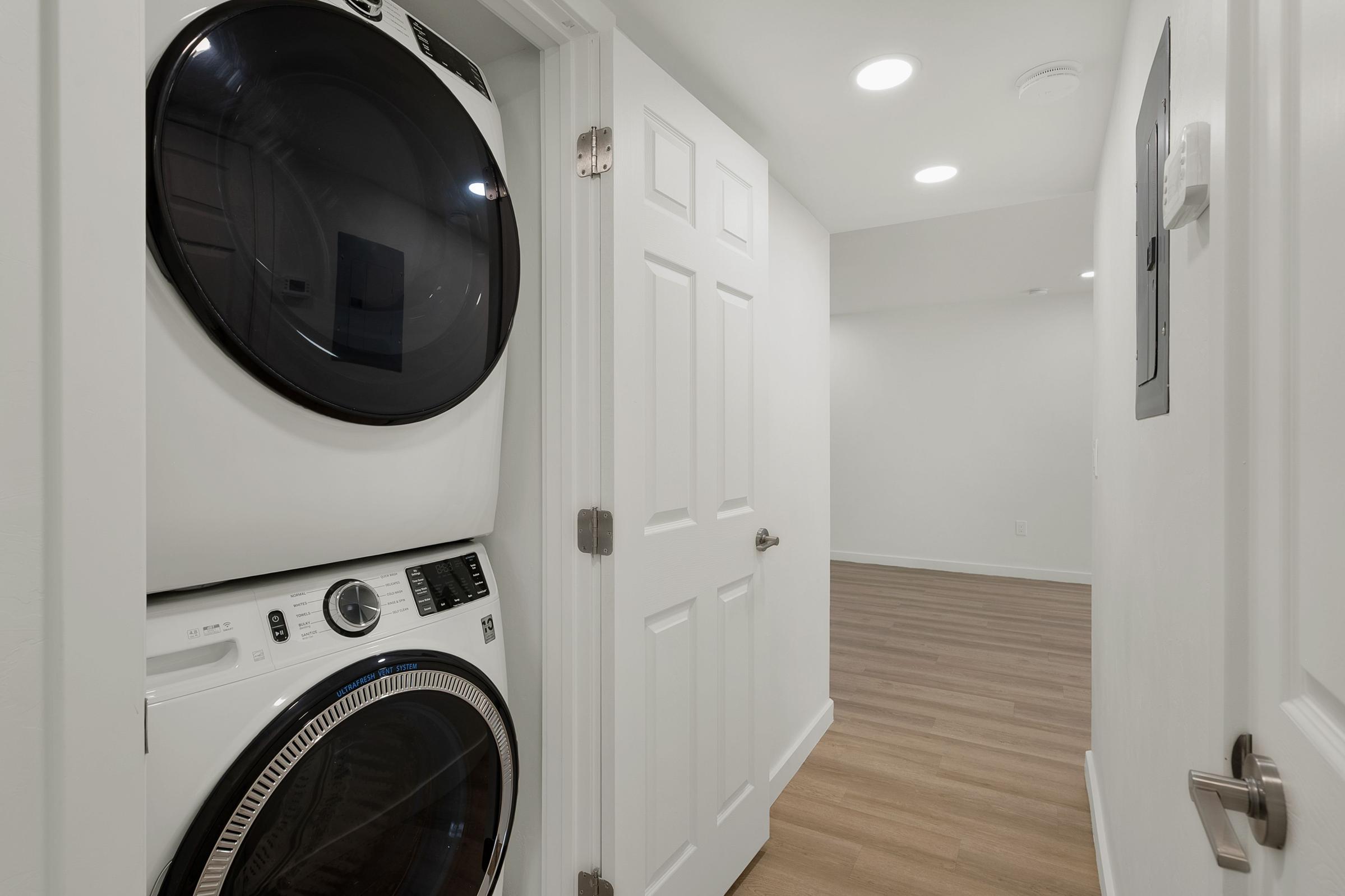 A compact laundry area featuring a stacked washer and dryer unit, located in a bright hallway with light-colored walls. The scene includes white doors leading to another room with wooden flooring and recessed ceiling lights, creating a modern and clean aesthetic.