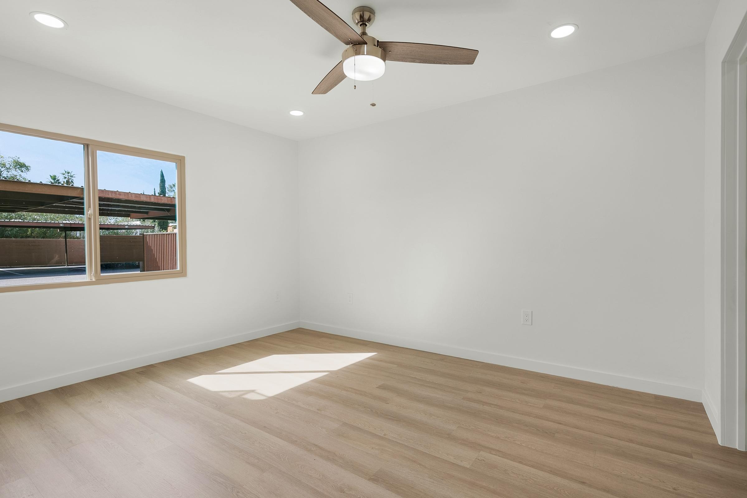 A spacious, well-lit room with light-colored walls and a wooden floor. It features a ceiling fan for air circulation and has a large window allowing natural light to enter. The view outside includes a patio area. The room is empty, providing a clean and minimalist aesthetic.