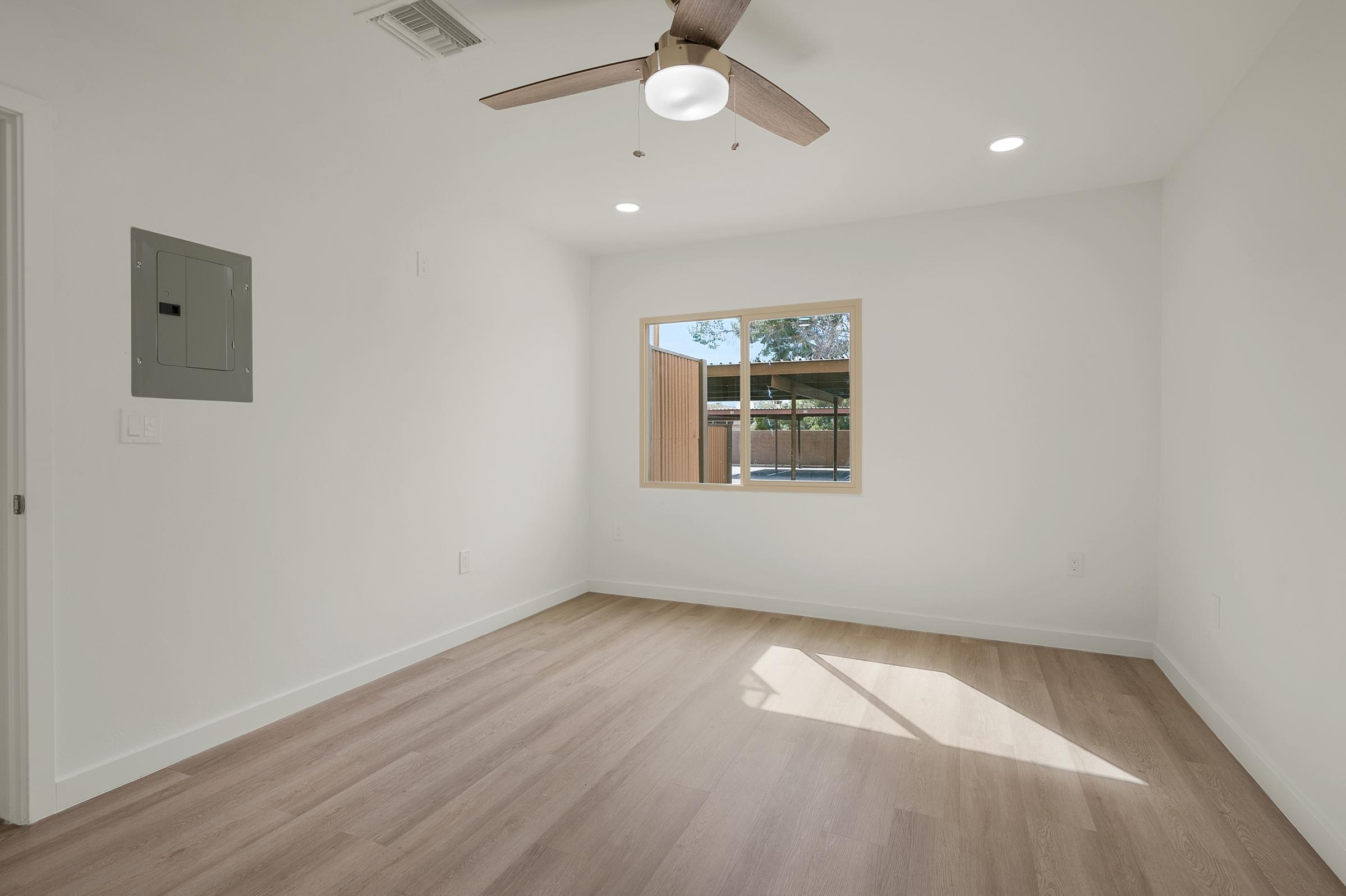 Empty room with light wood flooring, a ceiling fan, and natural light coming through a window. The walls are painted white, and a small electrical panel is visible on one wall. The space appears modern and well-lit, suitable for a living area or bedroom.