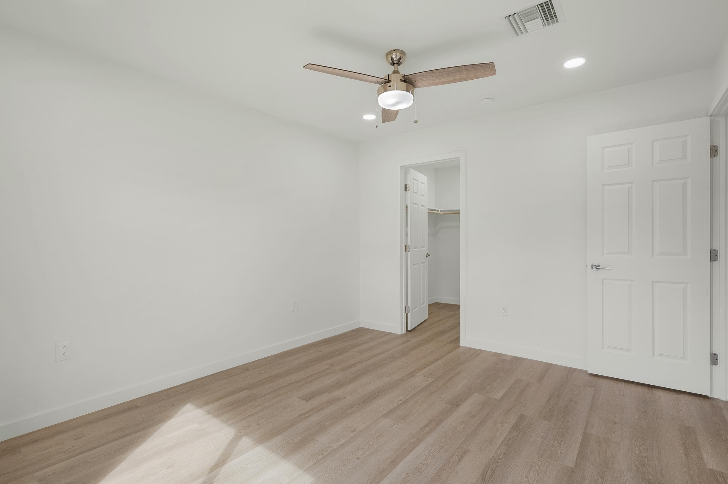 A bright, empty room with white walls and light wooden flooring. A ceiling fan with a light fixture hangs in the center. There's a closet visible in an adjacent room through an open white door. Natural light streams in, enhancing the spacious feel of the area.