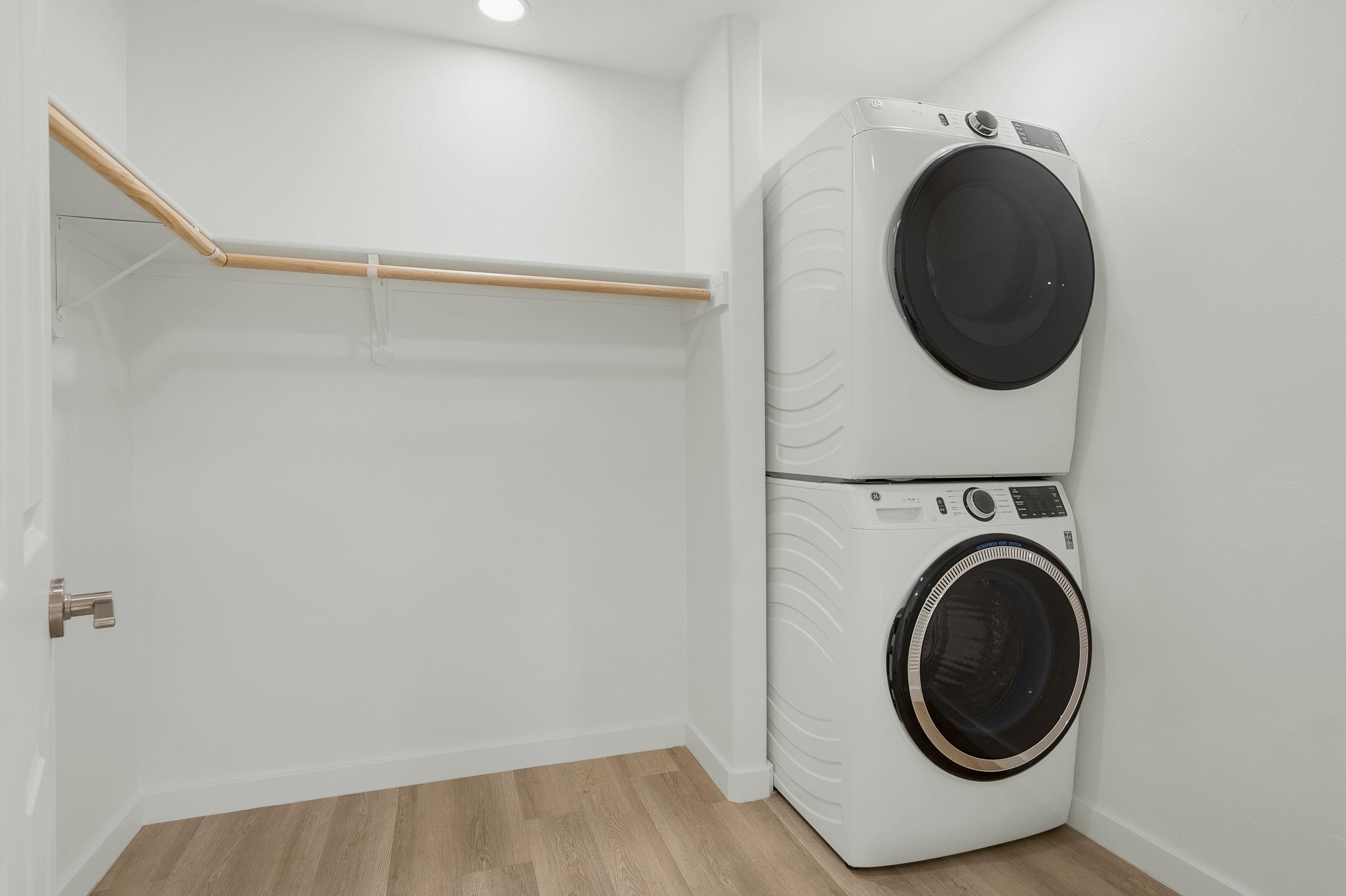 A modern laundry room featuring a stacked washer and dryer set in a light-colored space with wooden flooring. The room has a simple, clean design with a wooden hanging rod for storage and bright overhead lighting.