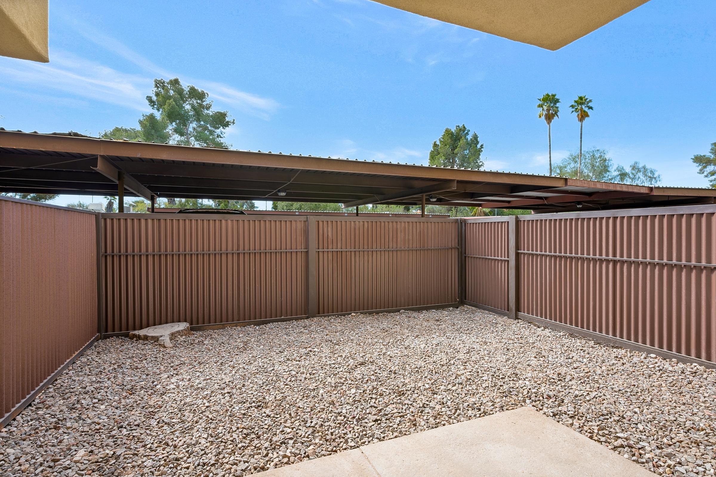 A small, fenced outdoor area featuring gravel flooring. The space is bordered by brown metal fencing and shaded by a roofed structure overhead. Palm trees are visible in the background against a clear blue sky, suggesting a warm, sunny environment.
