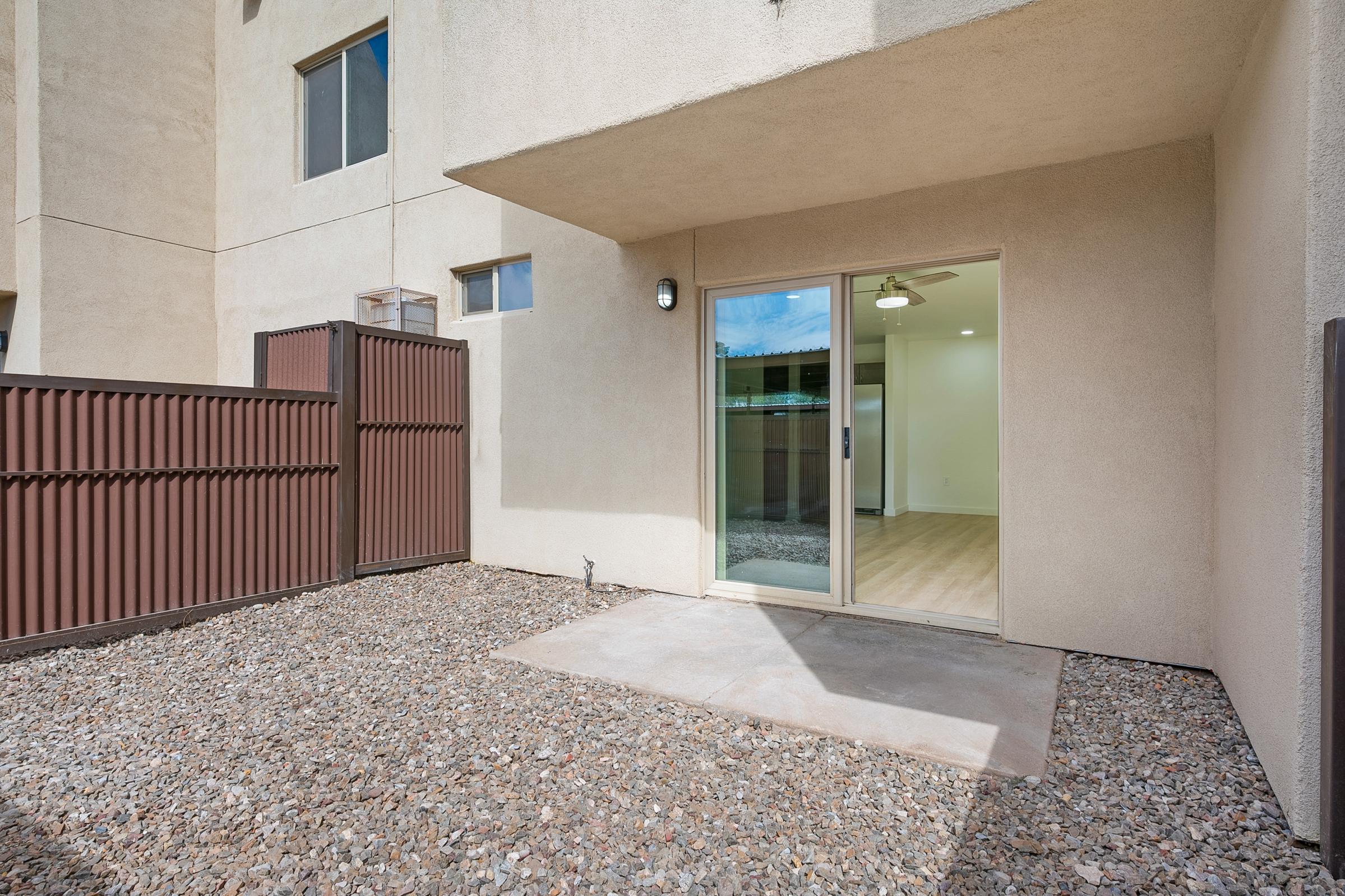 A small patio area with gravel flooring and a sliding glass door that leads inside from a beige-colored exterior wall. There is a brown fence on the side, and the interior space beyond the glass door features a ceiling fan and large windows.