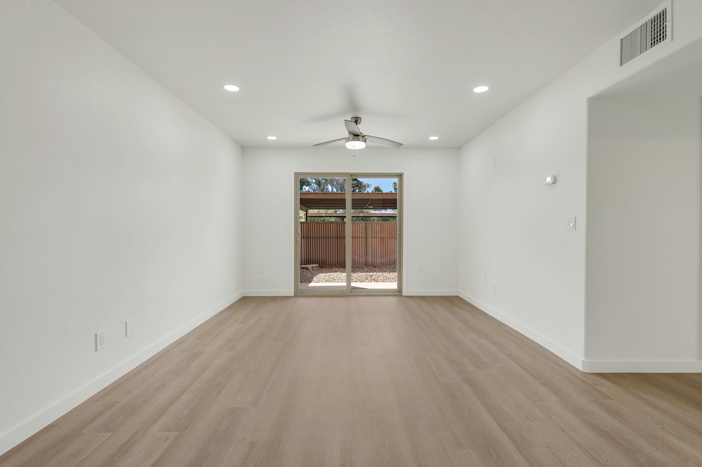 A spacious, empty interior room featuring light-colored walls and wooden flooring. There is a ceiling fan and large sliding glass doors that open to an outdoor area with a fenced yard, allowing natural light to enter the space. The room has a modern and minimalistic design.