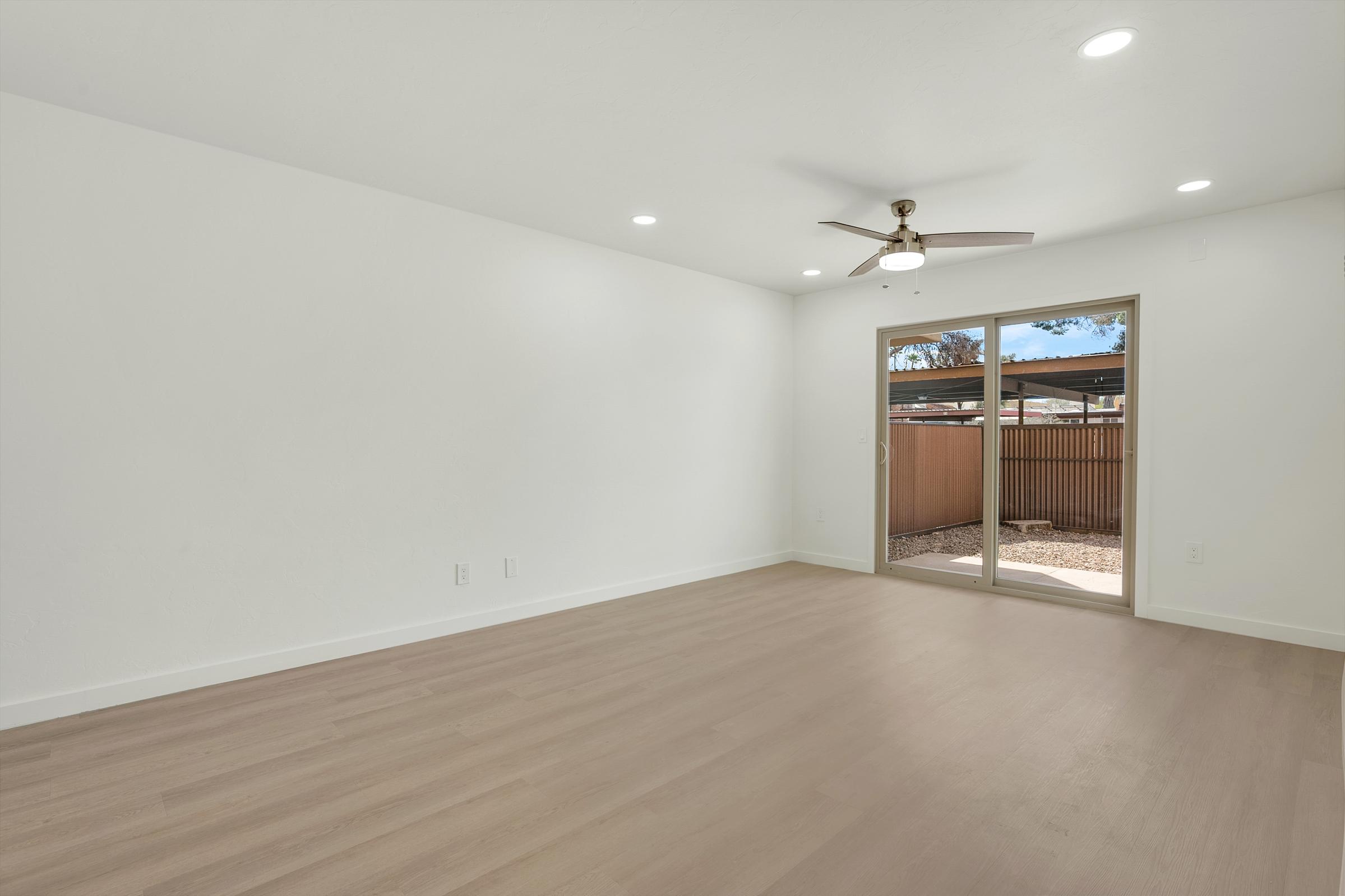 Empty interior of a room featuring light-colored walls, a ceiling fan, and large sliding glass doors leading to an outdoor space. The flooring is light wood, and the room is well-lit with natural light coming in through the doors.