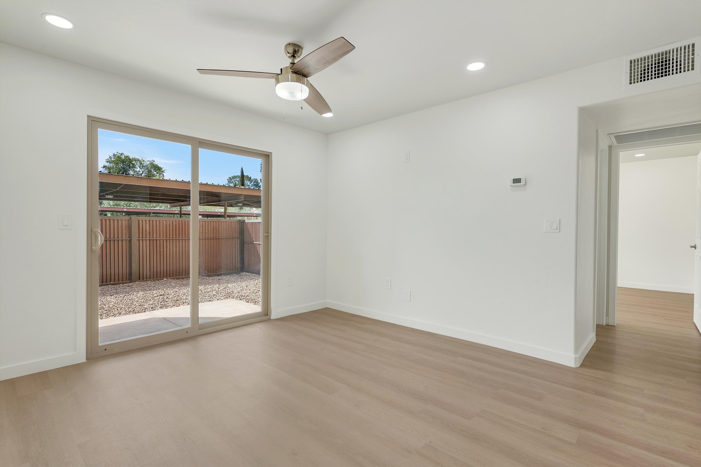 Bright, modern interior of a room with light wood flooring and white walls. A ceiling fan is mounted above, and large sliding glass doors provide a view of a fenced outdoor space. Natural light enters from the doors, enhancing the spacious feel. A hallway leads to another room, visible on the right.