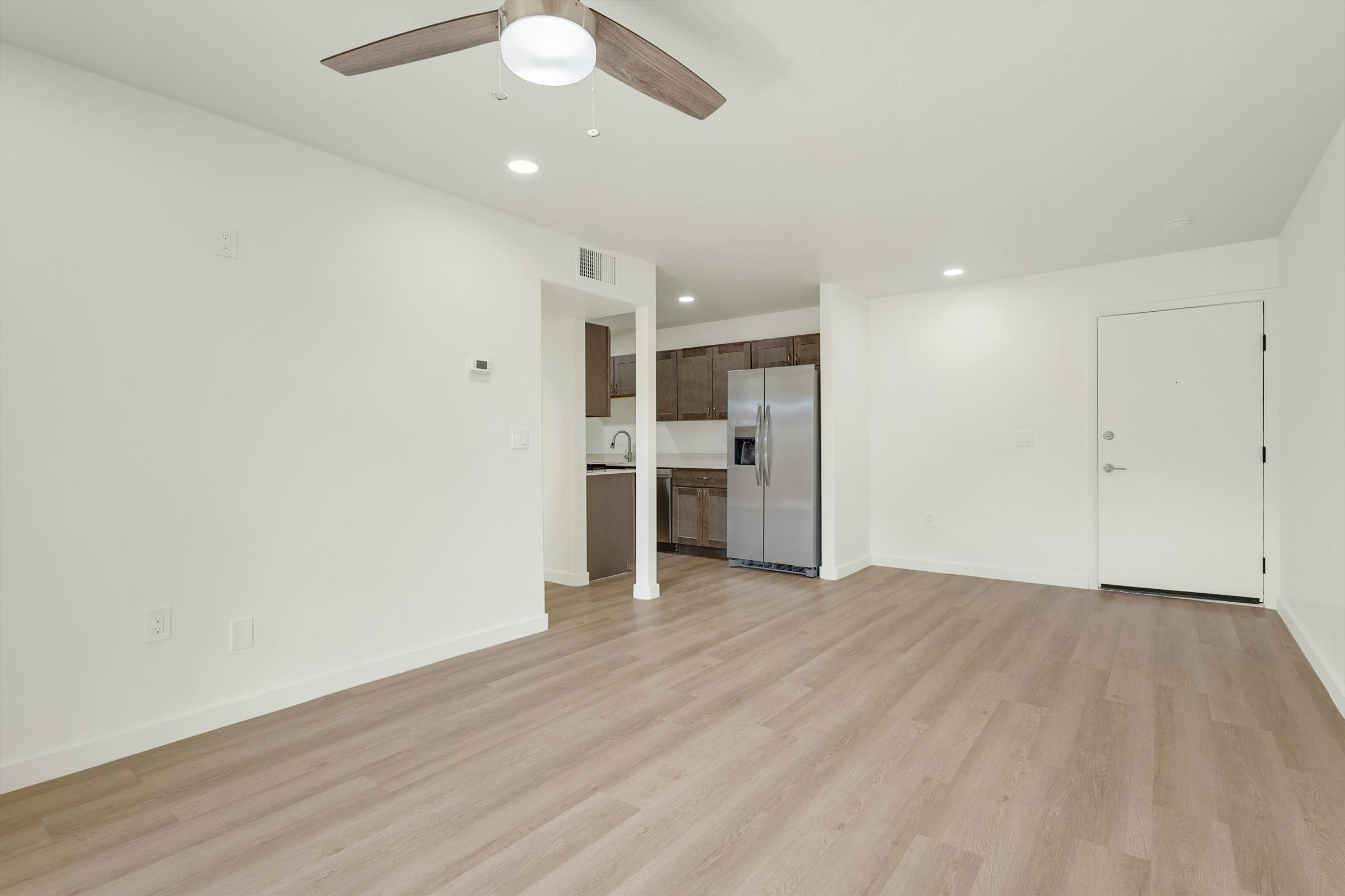 A modern, minimalist living space featuring light wood flooring, a ceiling fan, and an open layout. The kitchenette with dark wood cabinets and stainless steel appliances is visible in the background, along with a white door leading outside. The walls are painted in a bright, neutral color.