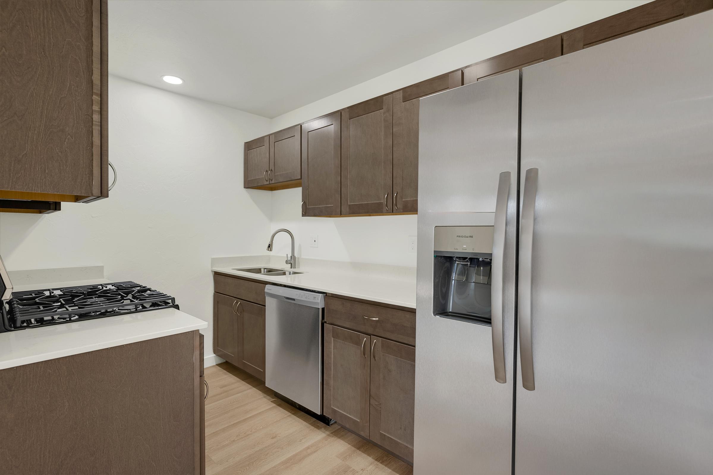Modern kitchen featuring stainless steel appliances, including a refrigerator and dishwasher, along with dark wood cabinetry. The countertop is light-colored, and the kitchen includes a gas stovetop. The space is well-lit with bright, neutral walls and hardwood flooring.