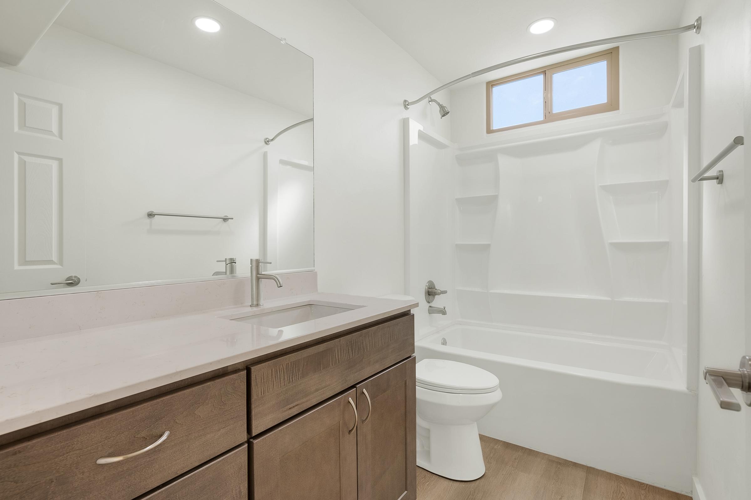 A modern bathroom featuring a clean design with a large mirror above a light-colored vanity, a white bathtub with built-in shelves, and a toilet. The room is well-lit by recessed ceiling lights and a small window allowing natural light. The flooring is light wood, creating a warm atmosphere.