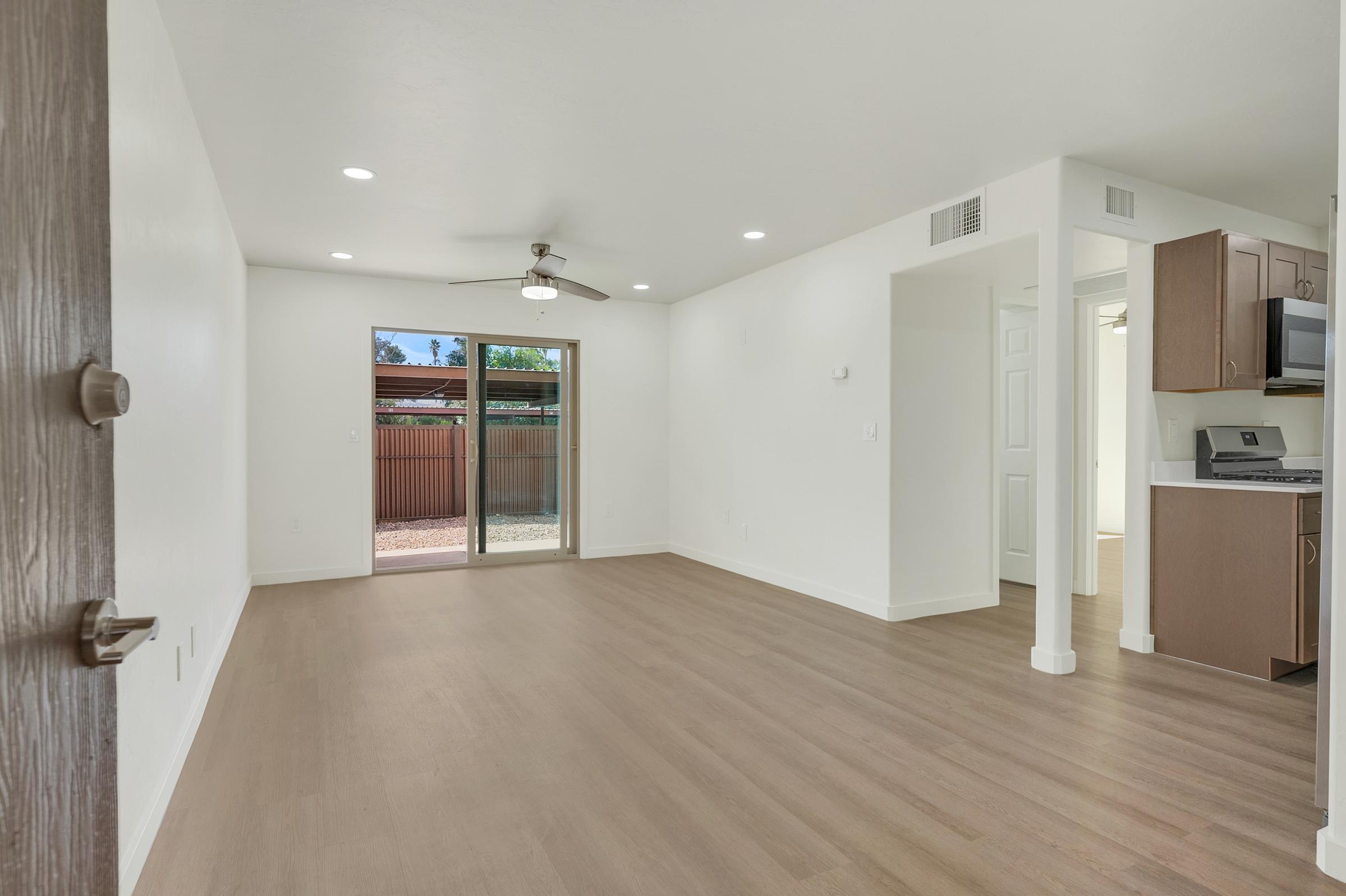 A spacious and bright living room with light hardwood flooring, white walls, and a ceiling fan. It features a sliding glass door leading to an outdoor area, with a view of a fenced yard. A kitchen area is visible on the right, showcasing modern cabinets and appliances. Natural light enhances the open layout.