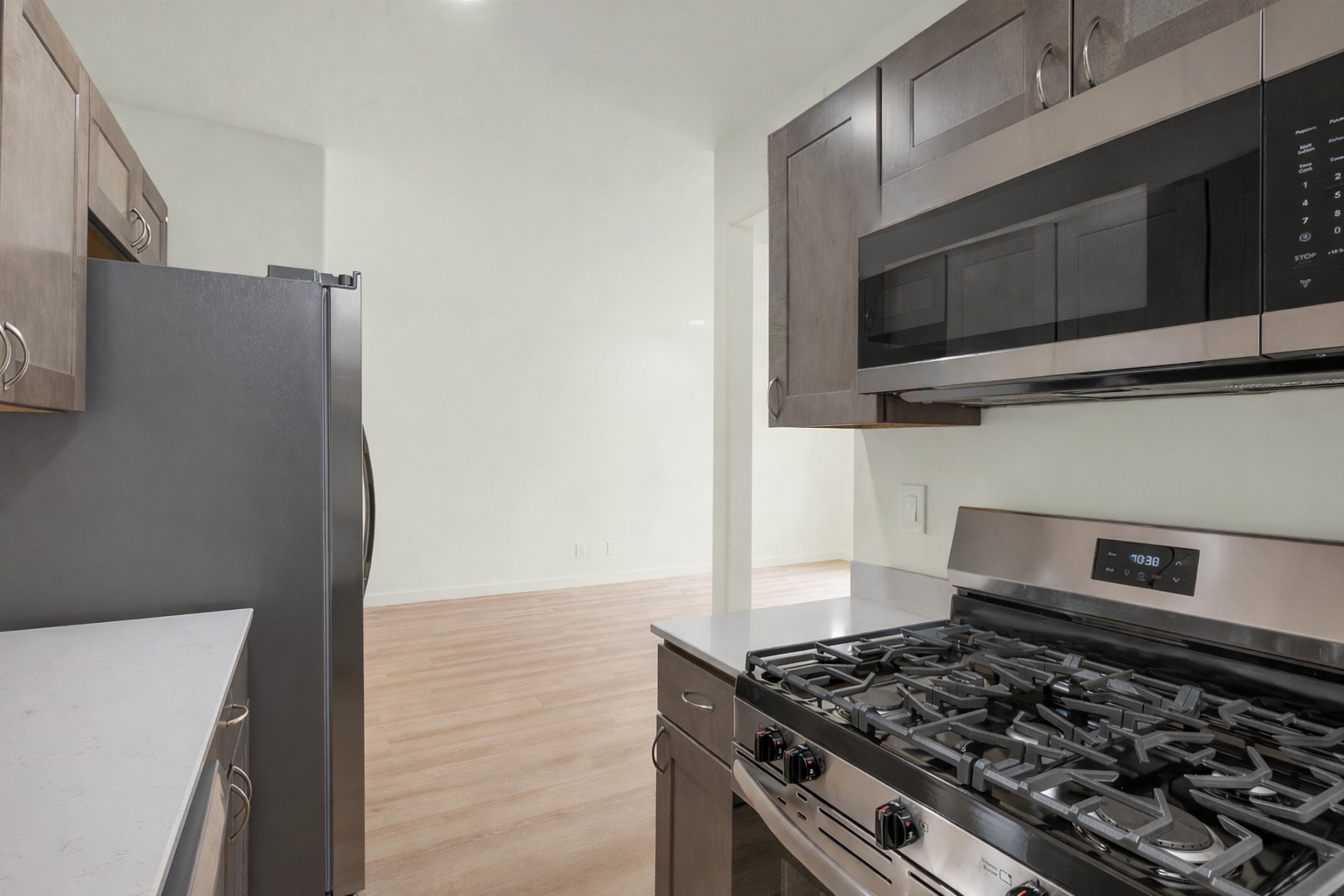 A modern kitchen featuring stainless steel appliances, including a gas stovetop and built-in microwave. The kitchen has wood cabinetry and a countertop with a light finish. A frosted glass door fridge is visible, and the background reveals a spacious, empty living area with light wood flooring.