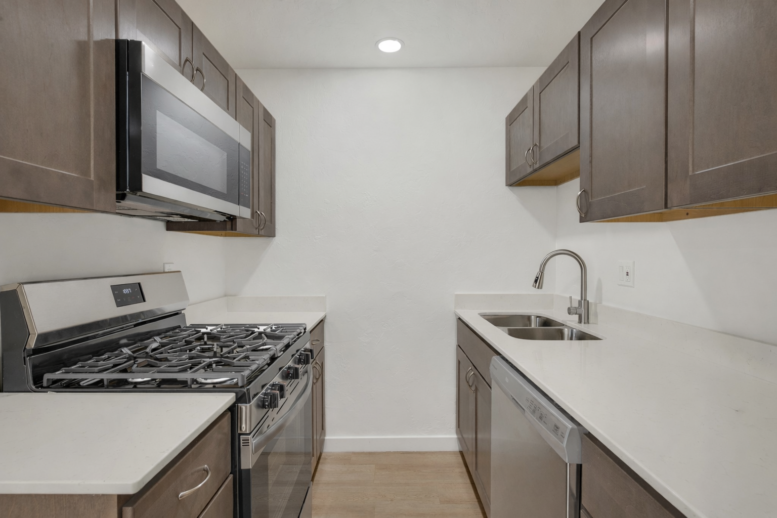 A modern kitchen featuring stainless steel appliances, including a gas stove, microwave, dishwasher, and a single basin sink. The cabinets are a dark wood finish, and the countertops are light-colored with a clean, minimalistic design. The walls are painted white, enhancing the bright and airy feel of the space.