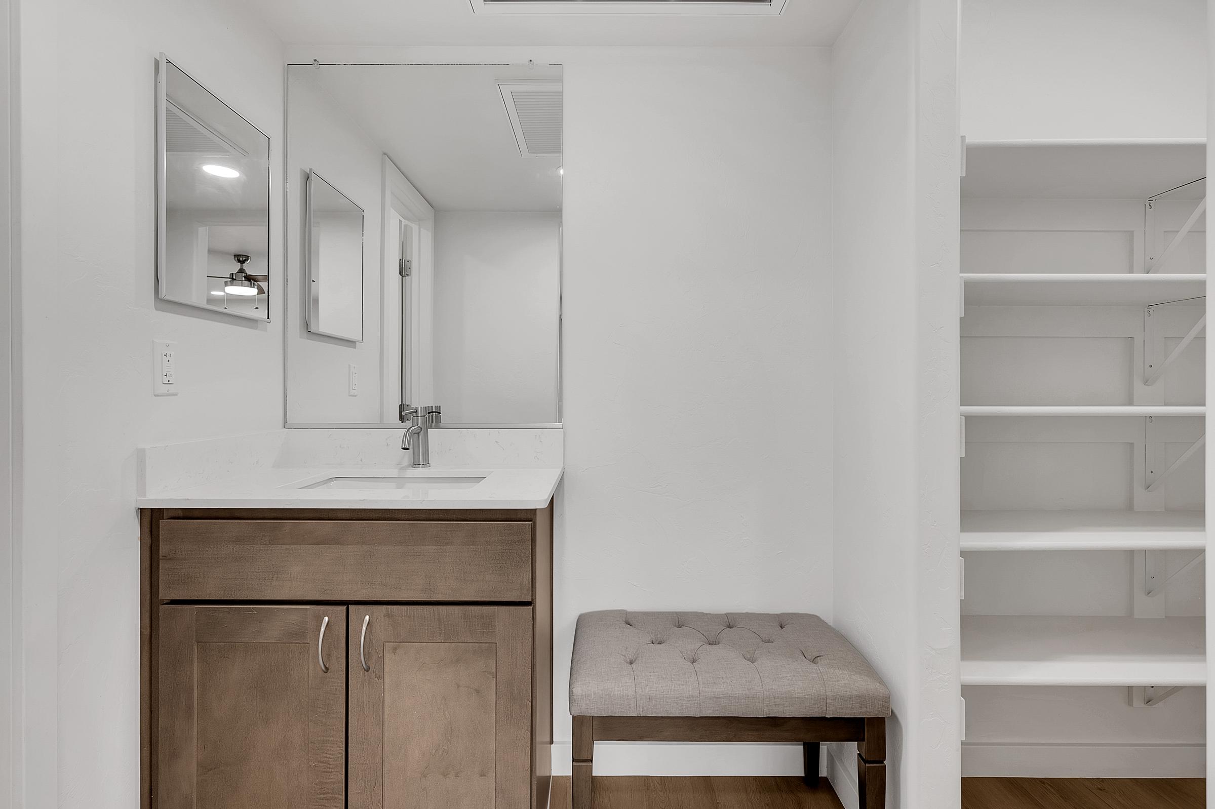 A clean, modern bathroom area featuring a white and gray color scheme. It includes a wooden vanity with a sink and a large mirror above it. There are two smaller mirrors on the wall, a cushioned bench in front of the vanity, and a white shelving unit to the right. The floor is light wood. 