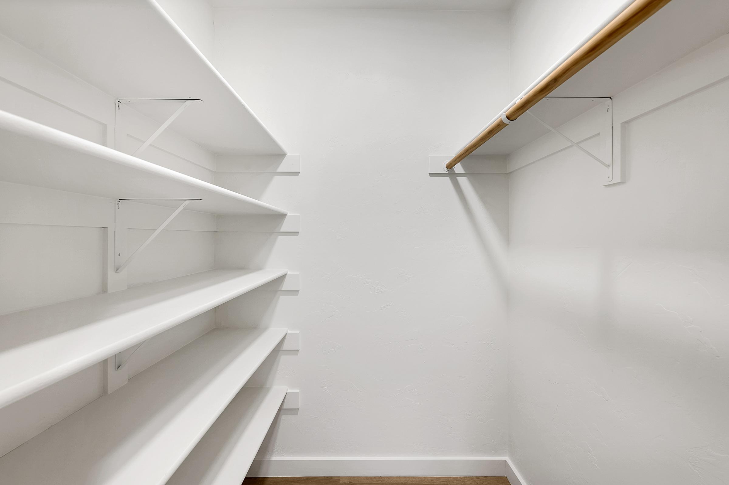 A bright and minimalistic closet with white walls and shelves. The closet features empty white shelves on the left side and a wooden rod for hanging clothes on the right, creating a clean and organized storage space. The floor is light-colored wood, enhancing the overall airy feel of the room.