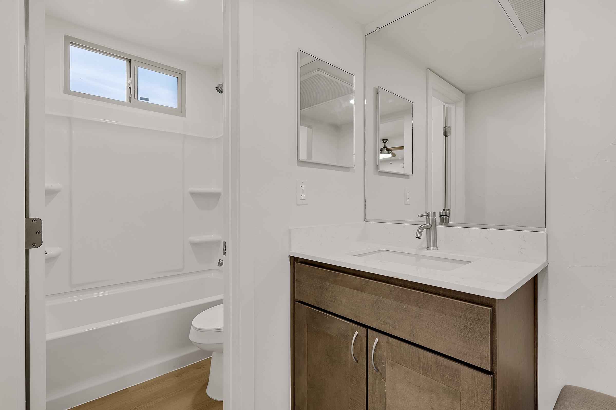 A modern bathroom featuring a white bathtub with a shower head, a wall-mounted mirror above a light-colored vanity with a sink, and a toilet. Natural light streams in through a small window, highlighting the clean lines and neutral color palette of the space.