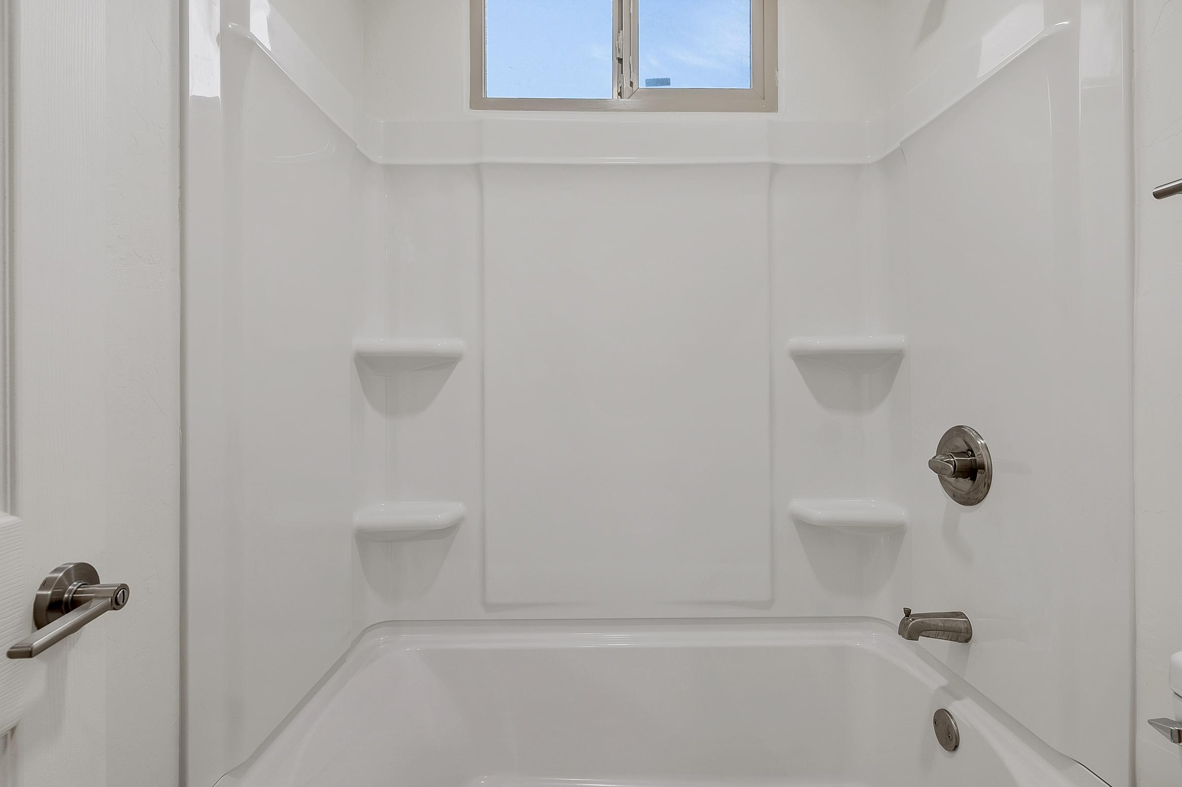 A clean and modern bathroom shower area featuring white walls, a bathtub, and built-in shelves. A small window above lets in natural light, and there are faucets and controls on the wall. The overall look is bright and minimalist.
