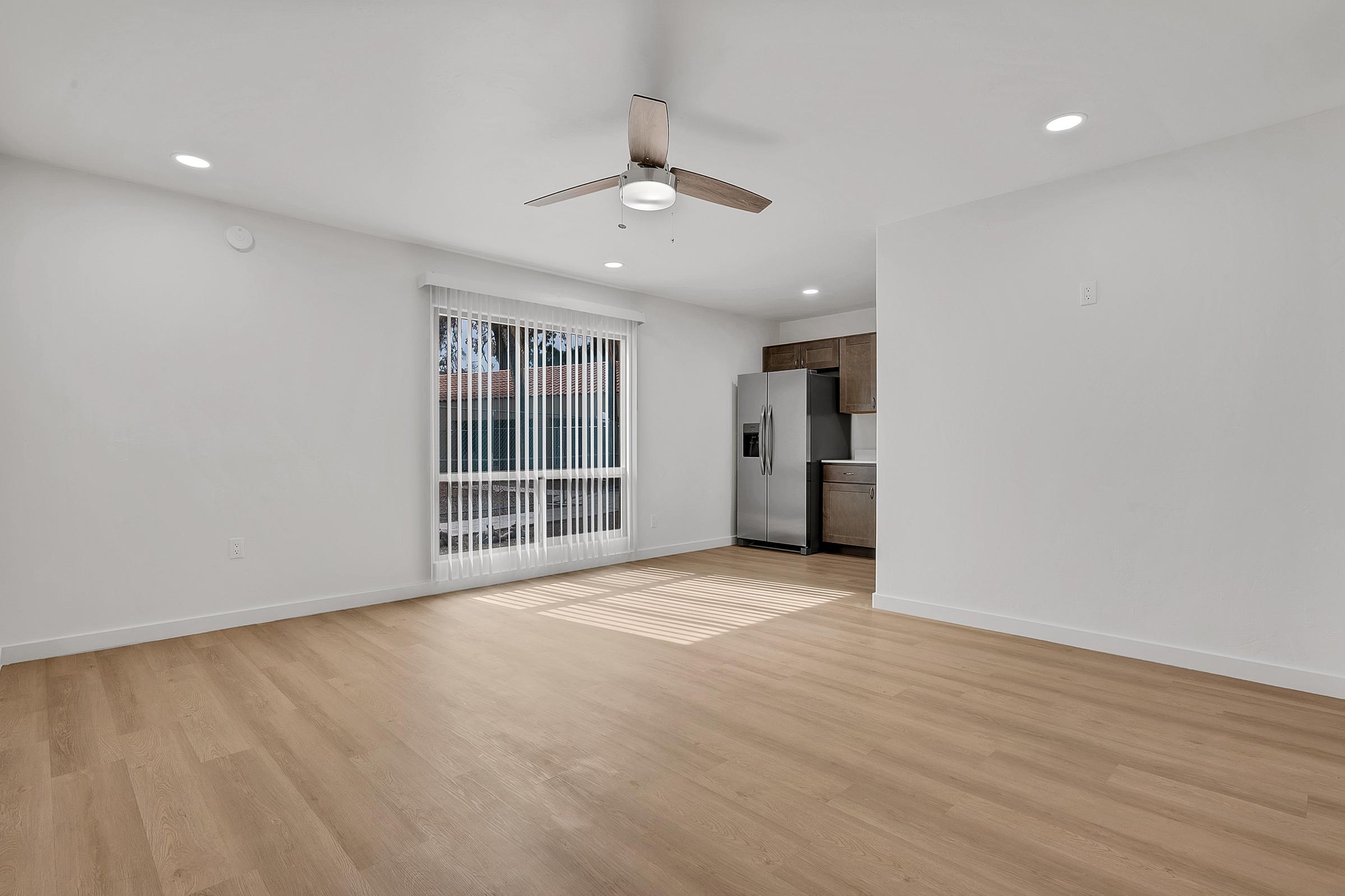 A bright, open living space with light-colored walls and wooden flooring. The room features a ceiling fan and large windows with vertical blinds. A modern kitchen area is visible in the background, showcasing a refrigerator and cabinetry. Natural light streams in, creating a spacious and inviting atmosphere.