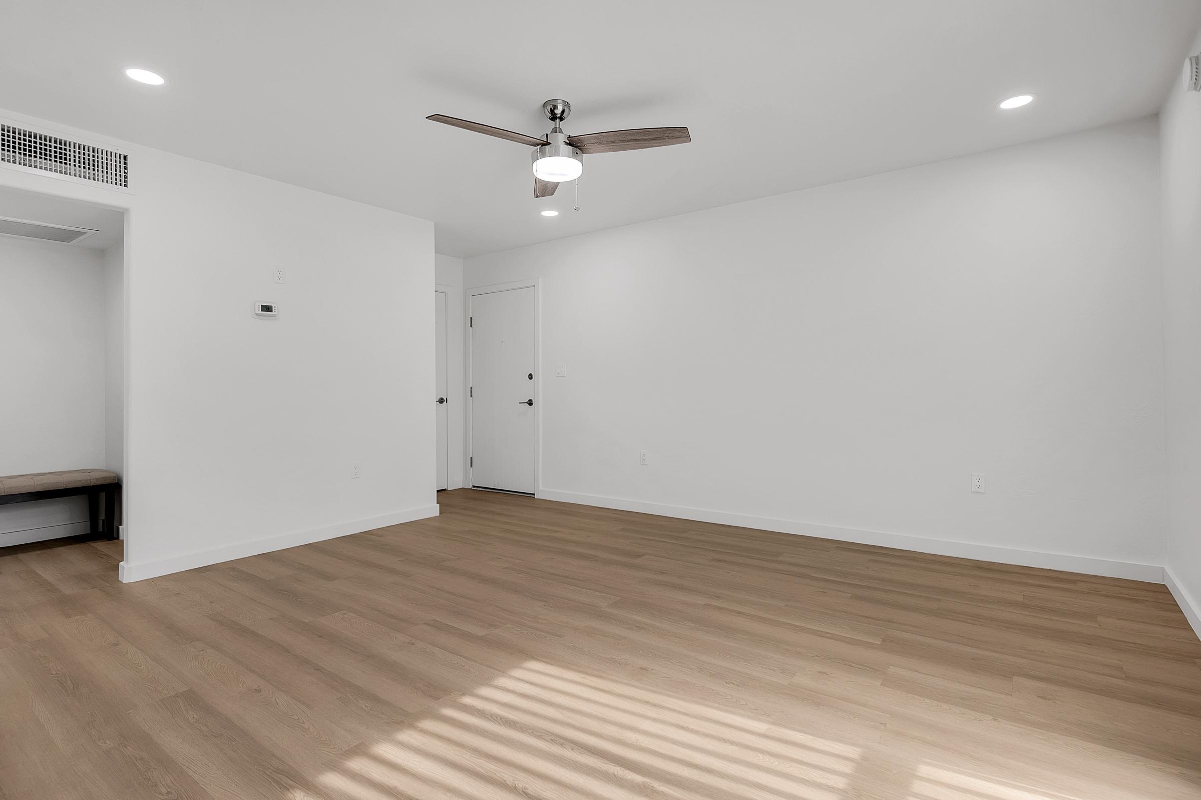 A bright, empty room with white walls and a wooden floor. It features a ceiling fan with a light fixture and has light streaming in through a window. A door leads to another space, and there is a small bench against the wall near the entrance. The room has a minimalist design, creating a spacious feel.